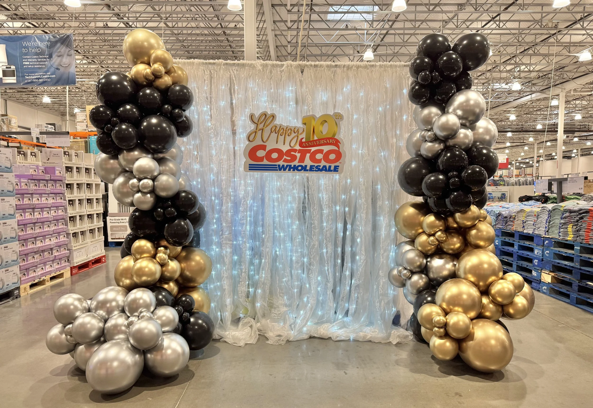 Balloon arch with black, silver, and gold balloons surrounding a backdrop with a sign that reads 'Happy 10th Anniversary Costco Wholesale,' decorated with white lights in a retail store. Corporate Events