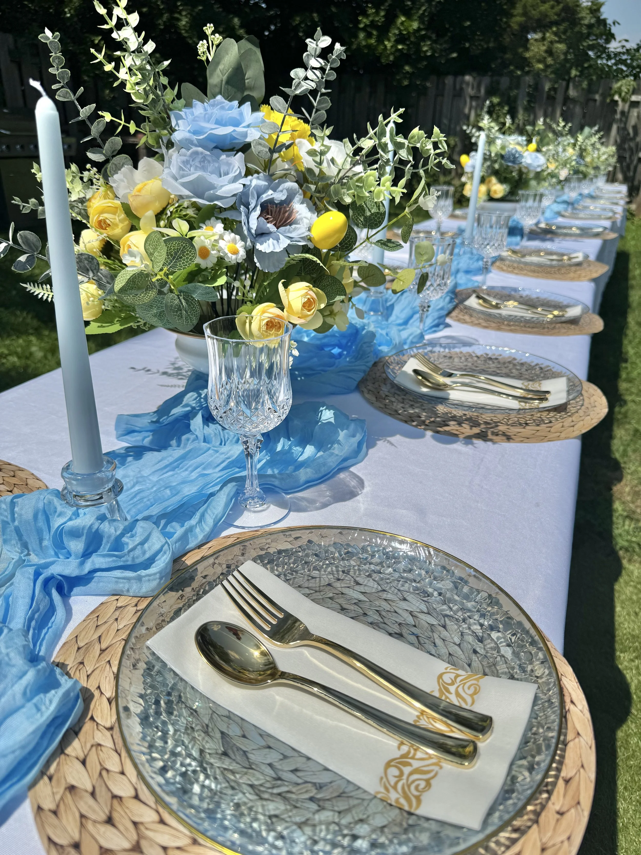 A long table set outdoors with a white tablecloth, decorated with a blue runner, a bouquet of blue, yellow, and white flowers, gold cutlery, glasses, and woven placemats, with sunlight and a wooden fence in the background.