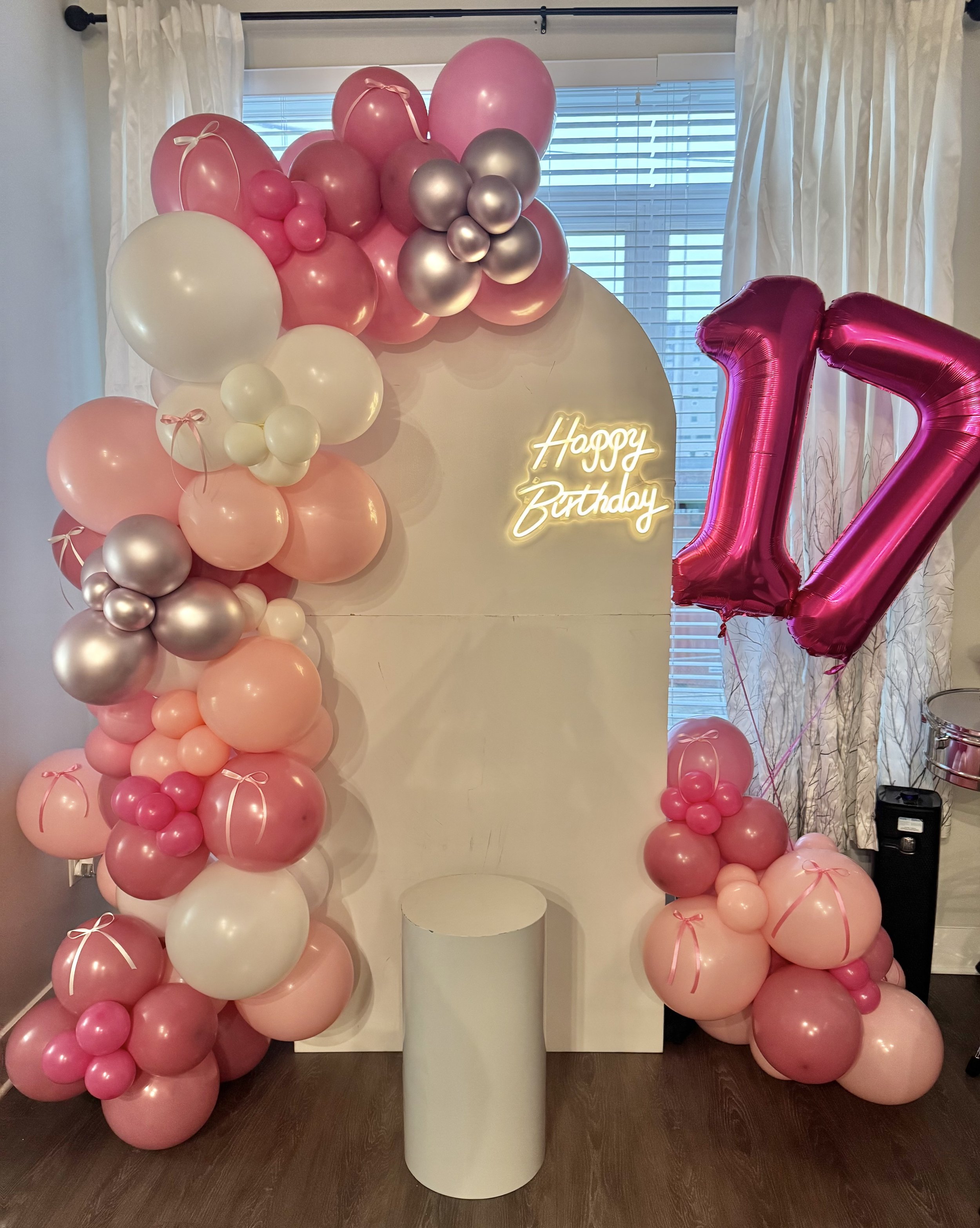 A birthday celebration setup with a balloon arch, large pink number 12 balloon, a neon 'Happy Birthday' sign, and a white backdrop with curtains in the background.