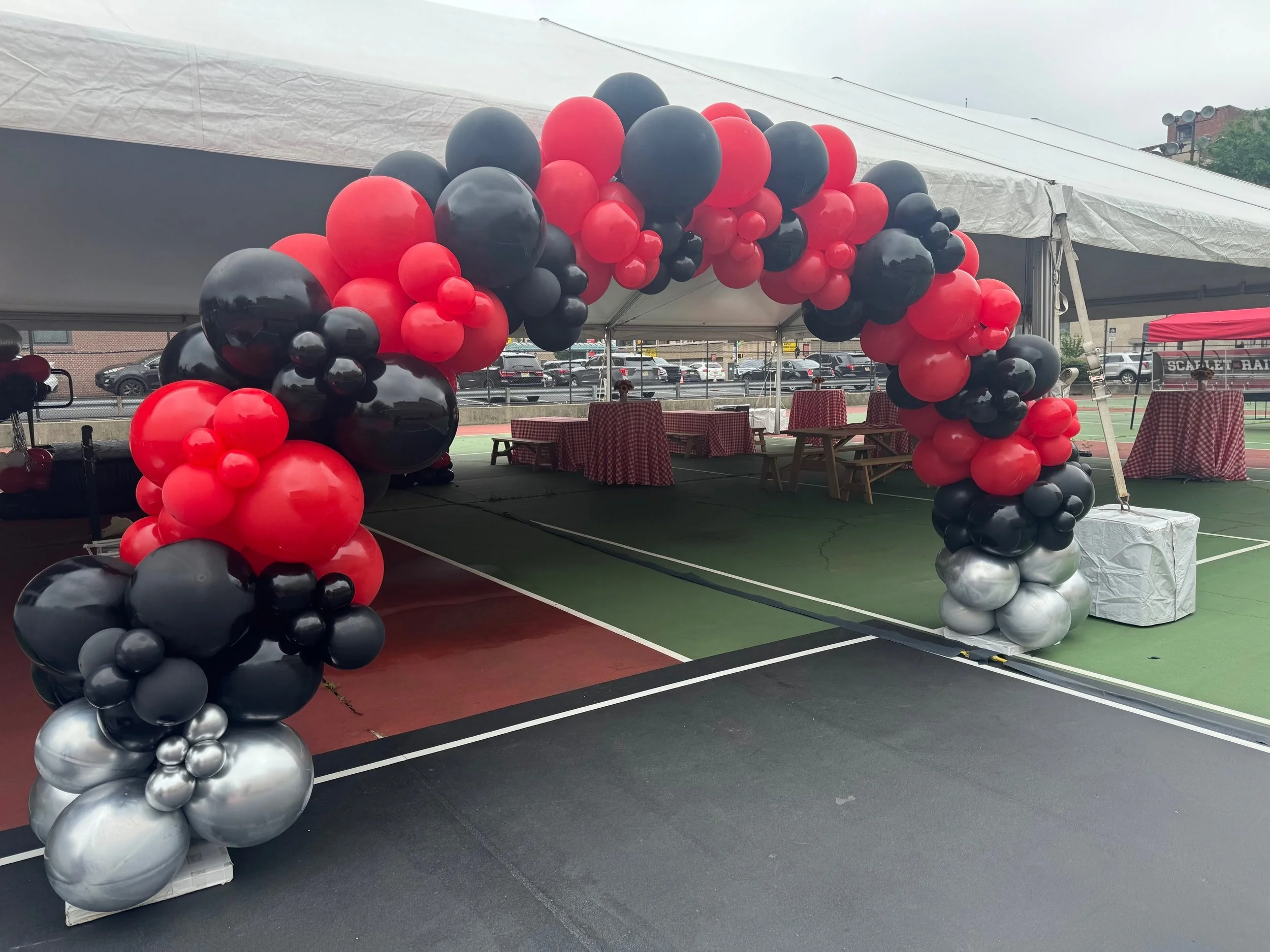 Balloon arch with black, red, and silver balloons set up on a sports court under a white canopy tent.