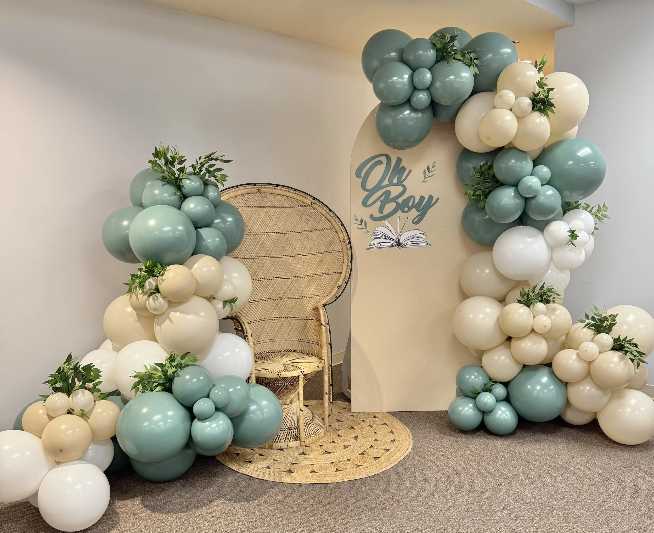 Balloon arch in shades of mint green, white, and cream, decorated with greenery, with a wedding or celebration backdrop sign reading 'Oh Boy' and an open book illustration.