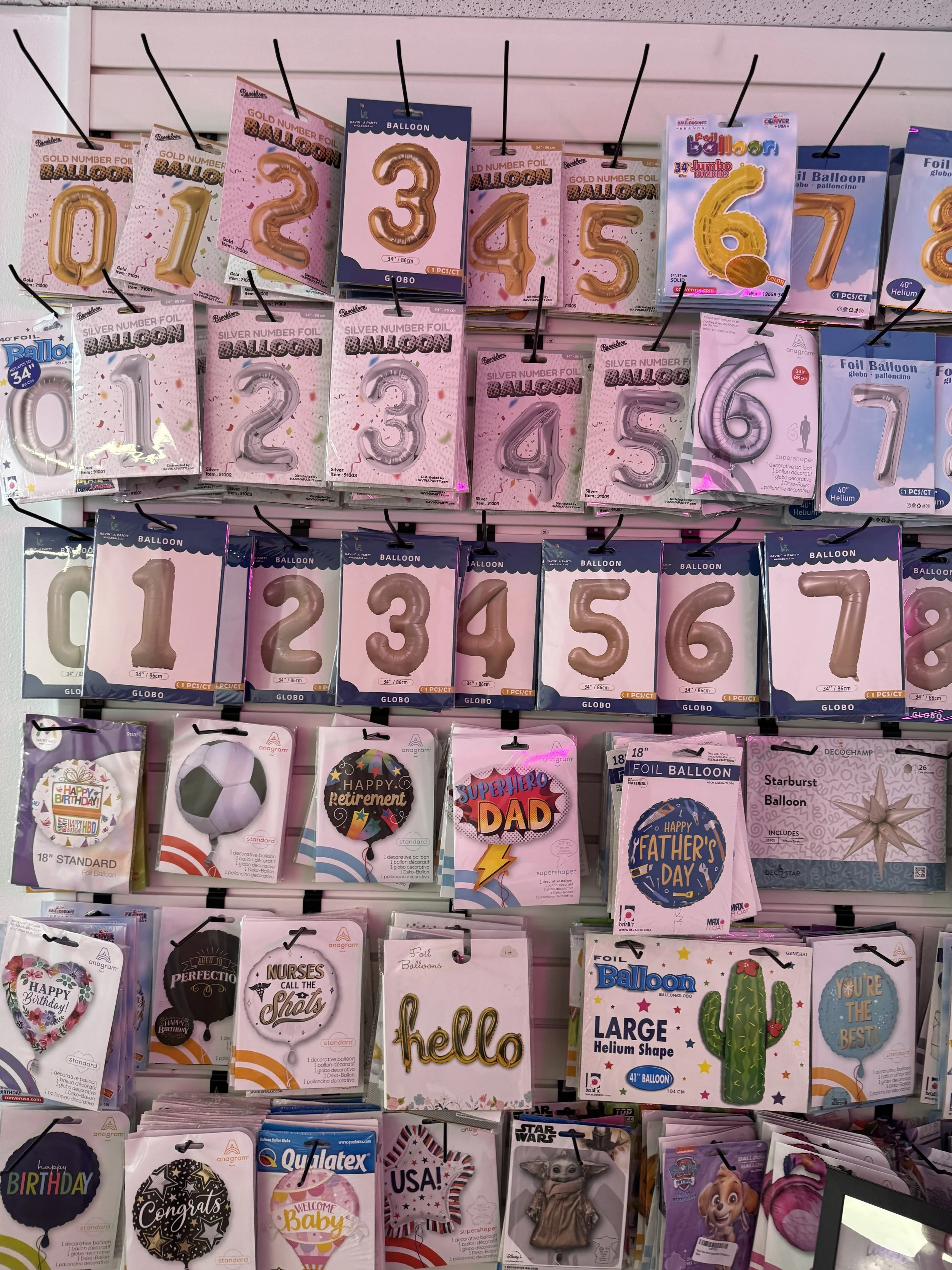 Display of various foil balloons in the shapes of numbers from 0 to 9, some with additional designs or phrases, arranged on hooks on a store wall.