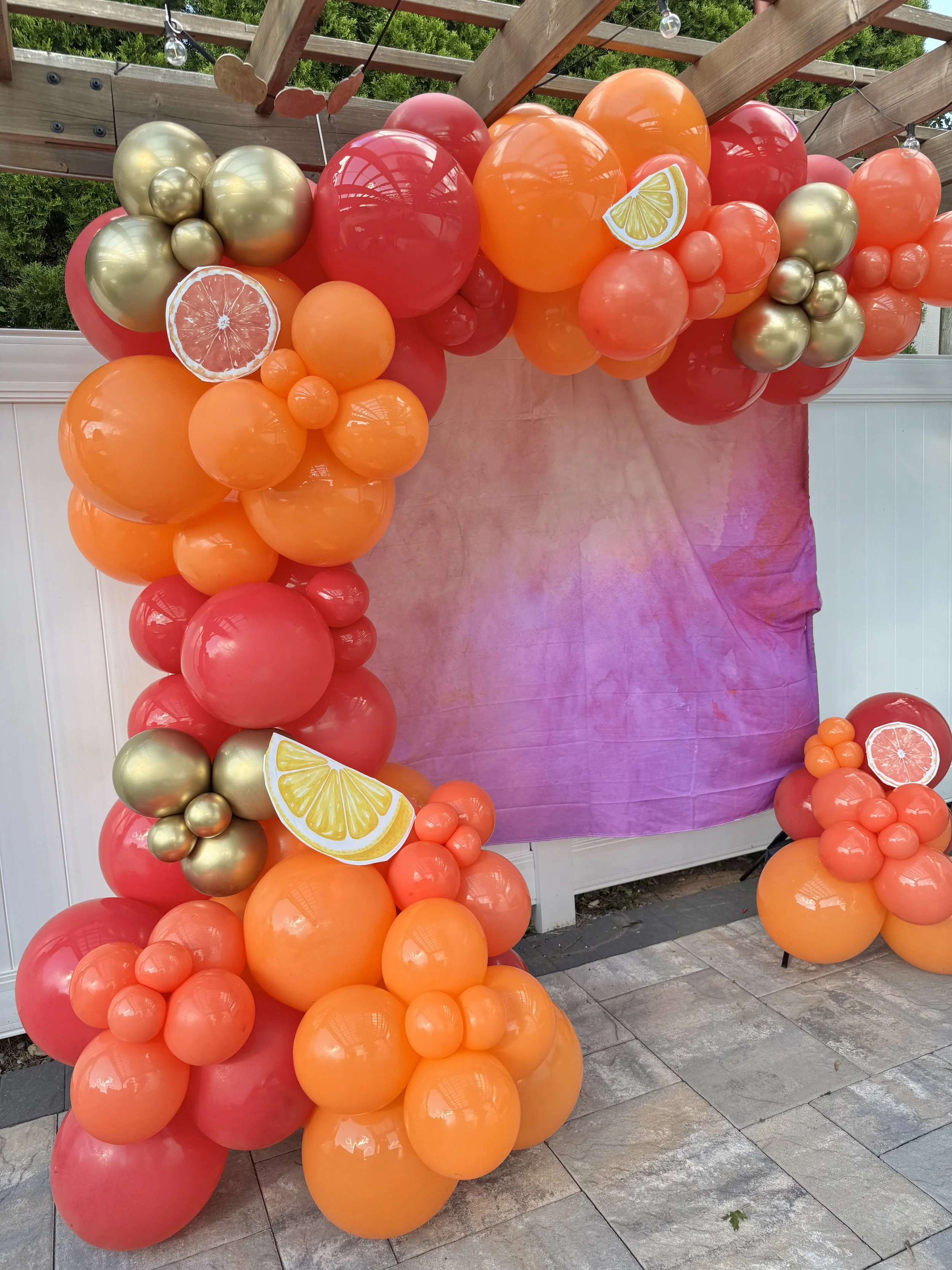 Decorative balloon arch with red, orange, gold, and pink balloons, adorned with paper lemon and grapefruit slices, set against a pink backdrop.