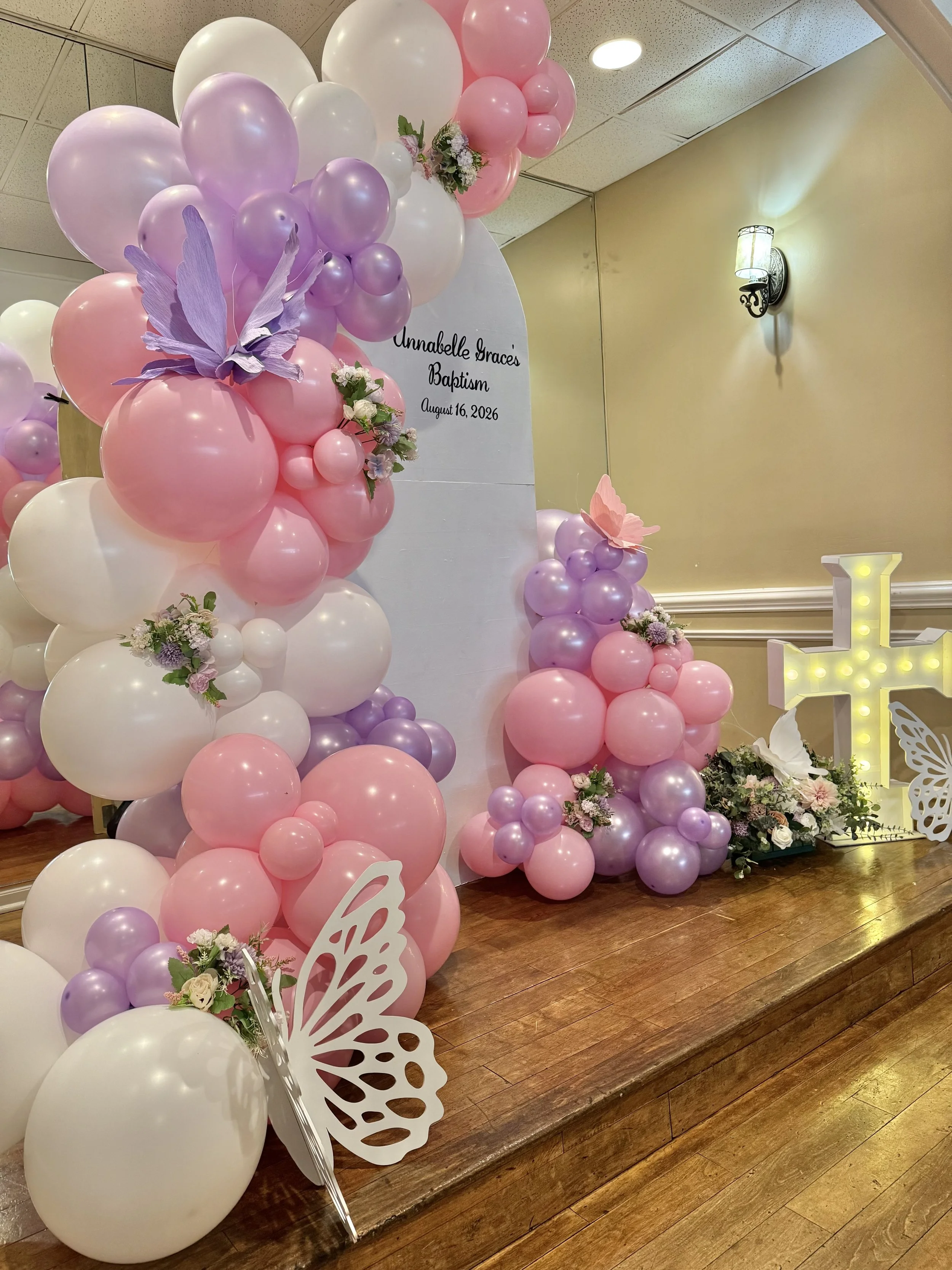 Decorative setup with pink, purple, and white balloons arranged around a white backdrop with flowers and butterfly cutouts for a baptism celebration. Sign reads 'Annabelle Grace's Baptism August 16, 2026'.