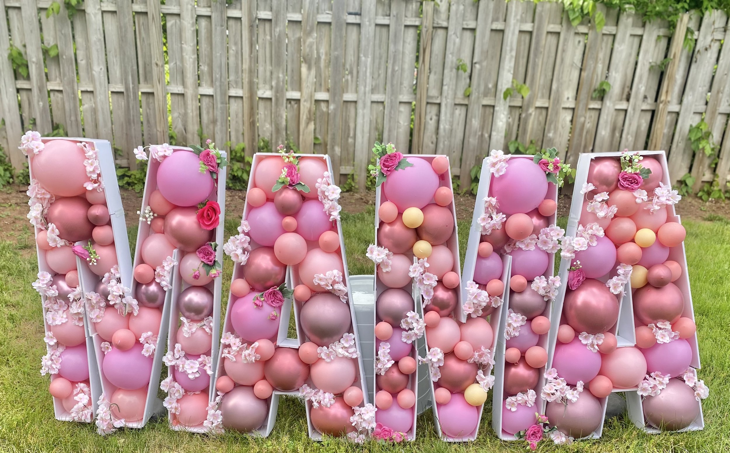 Large decorative letter spelling 'WALM' made of pink and rose gold balloons, decorated with pink flowers, placed outdoors on grass with a wooden fence background.
