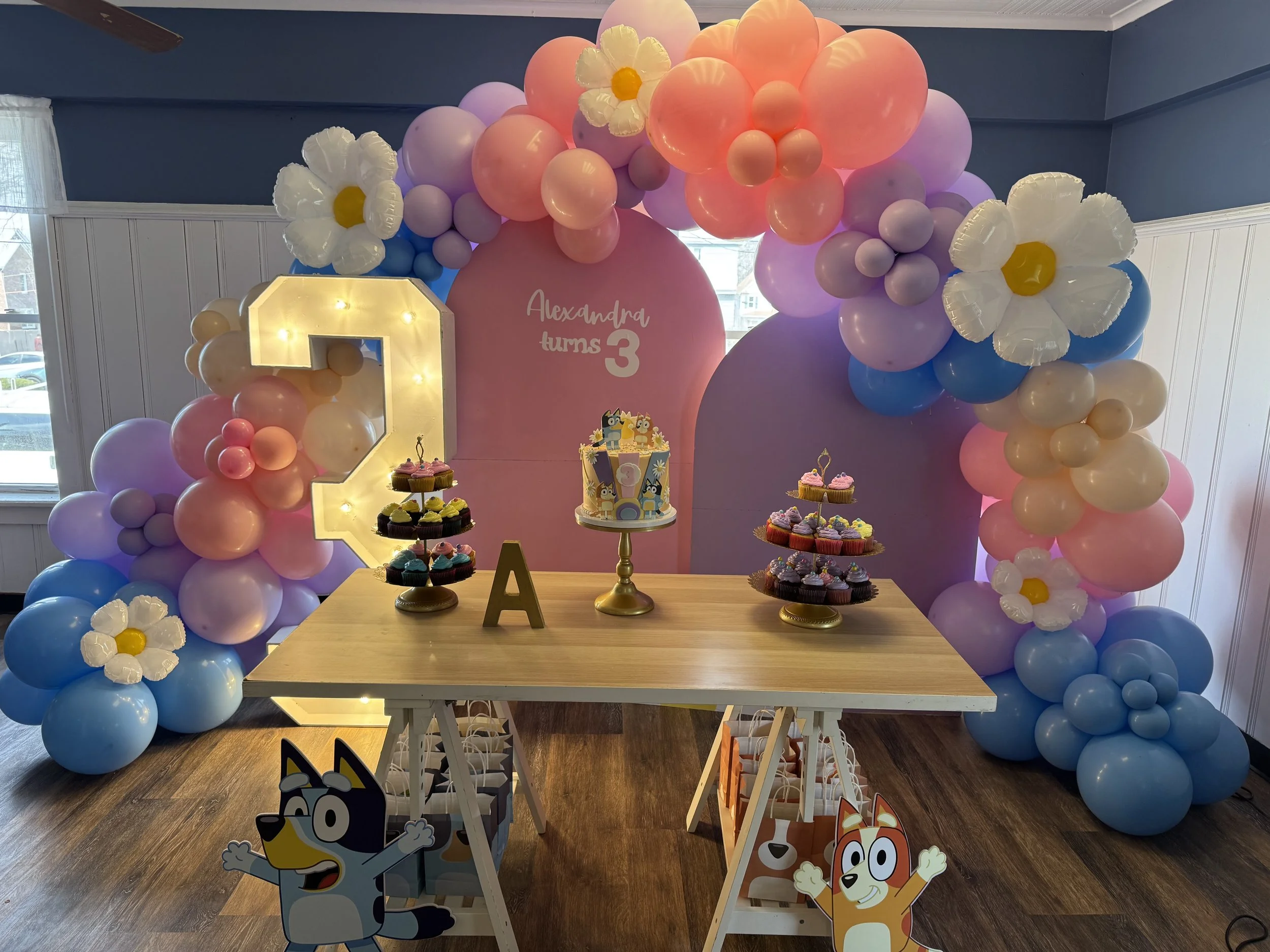 Colorful balloon arch with purple, pink, blue, and white balloons, decorated with large paper flowers, framing a pink backdrop that reads 'Alexandra turns 3.' A table with cupcakes and a cake featuring cartoon characters is in front, with animated ch