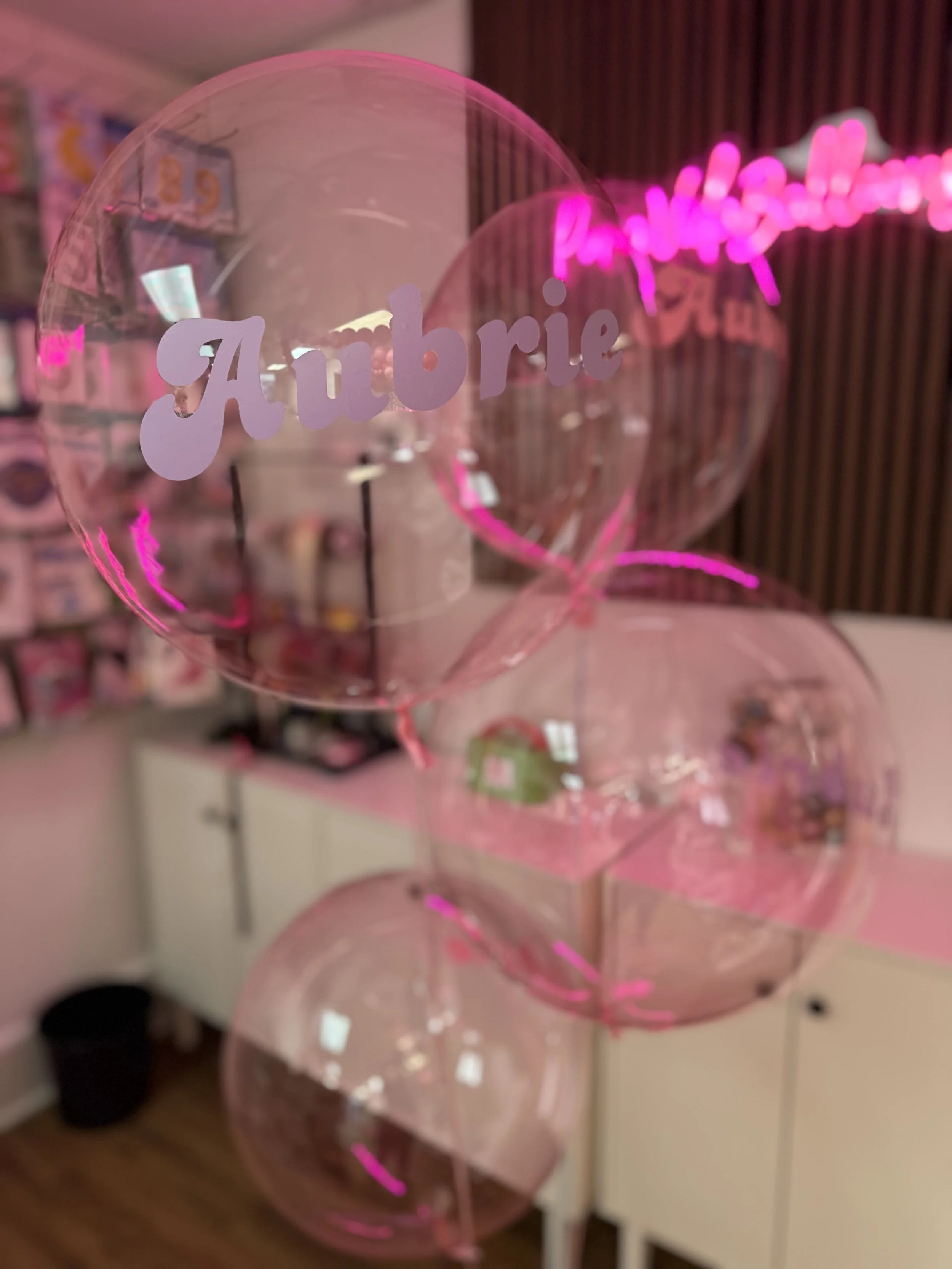 Pink transparent balloons with the word 'Ambie' written on one balloon, set in a room with a pink neon sign in the background.