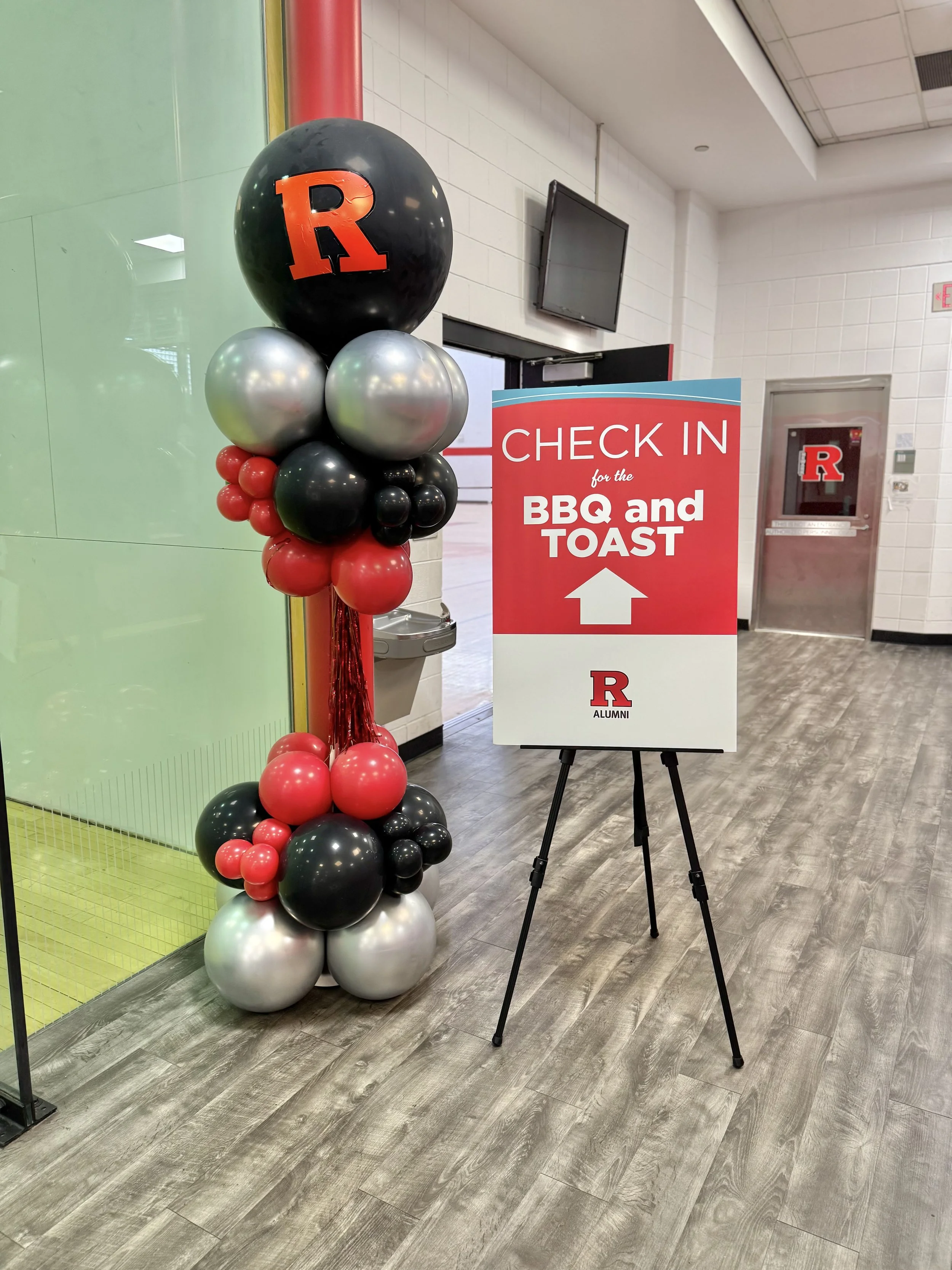 A decor of balloons and a sign in a hallway. The balloons are arranged in a pillar with black, silver, red, and white colors, featuring a large black balloon with a red 'R'. The sign indicates check-in for the university's BBQ and Toast event, with a