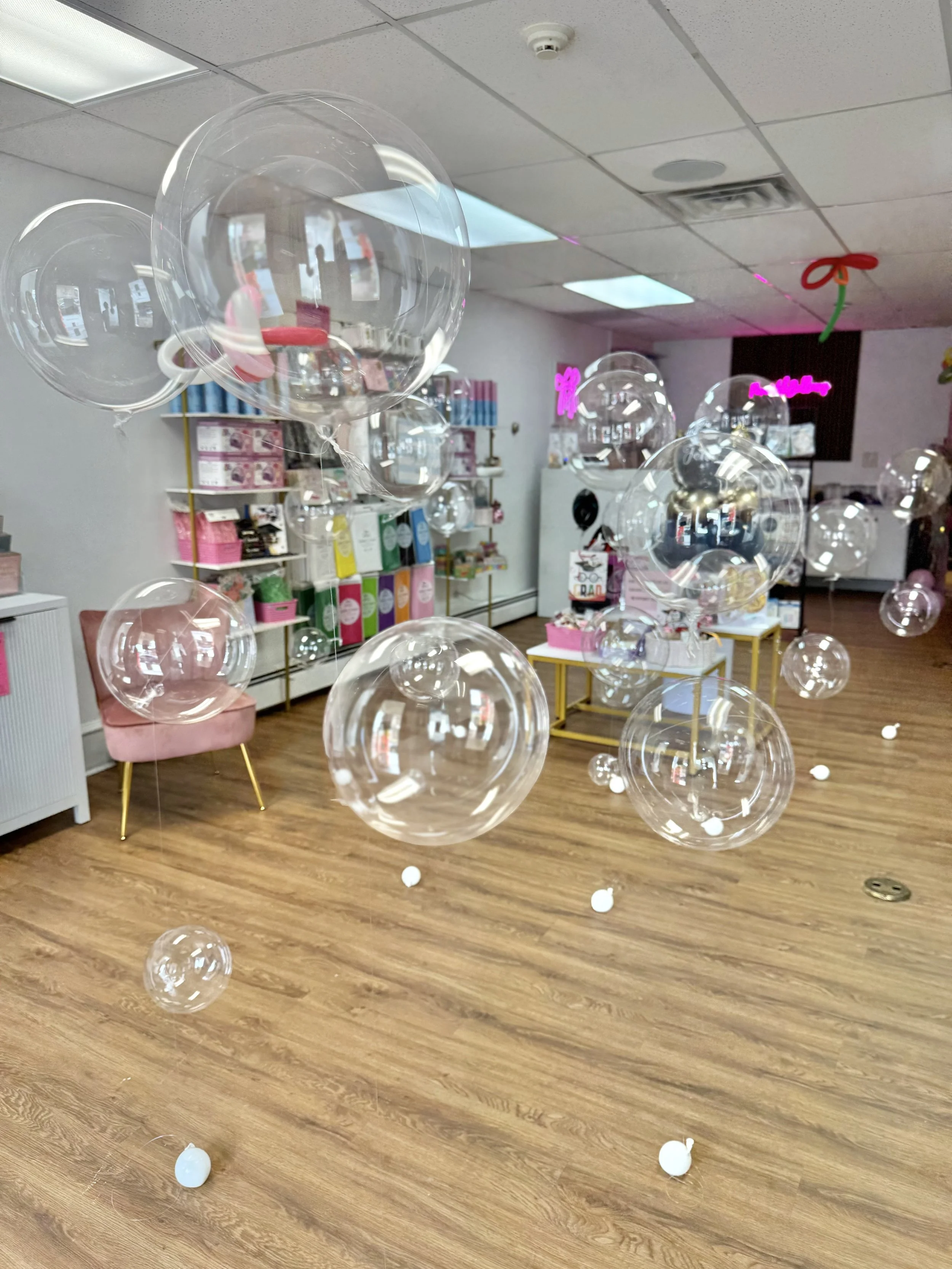 A room with a ceiling of tiles and wood flooring. There are see-through balloons floating and some on the floor. In the background, shelves hold colorful boxes and other items, and neon signs are on the back wall.