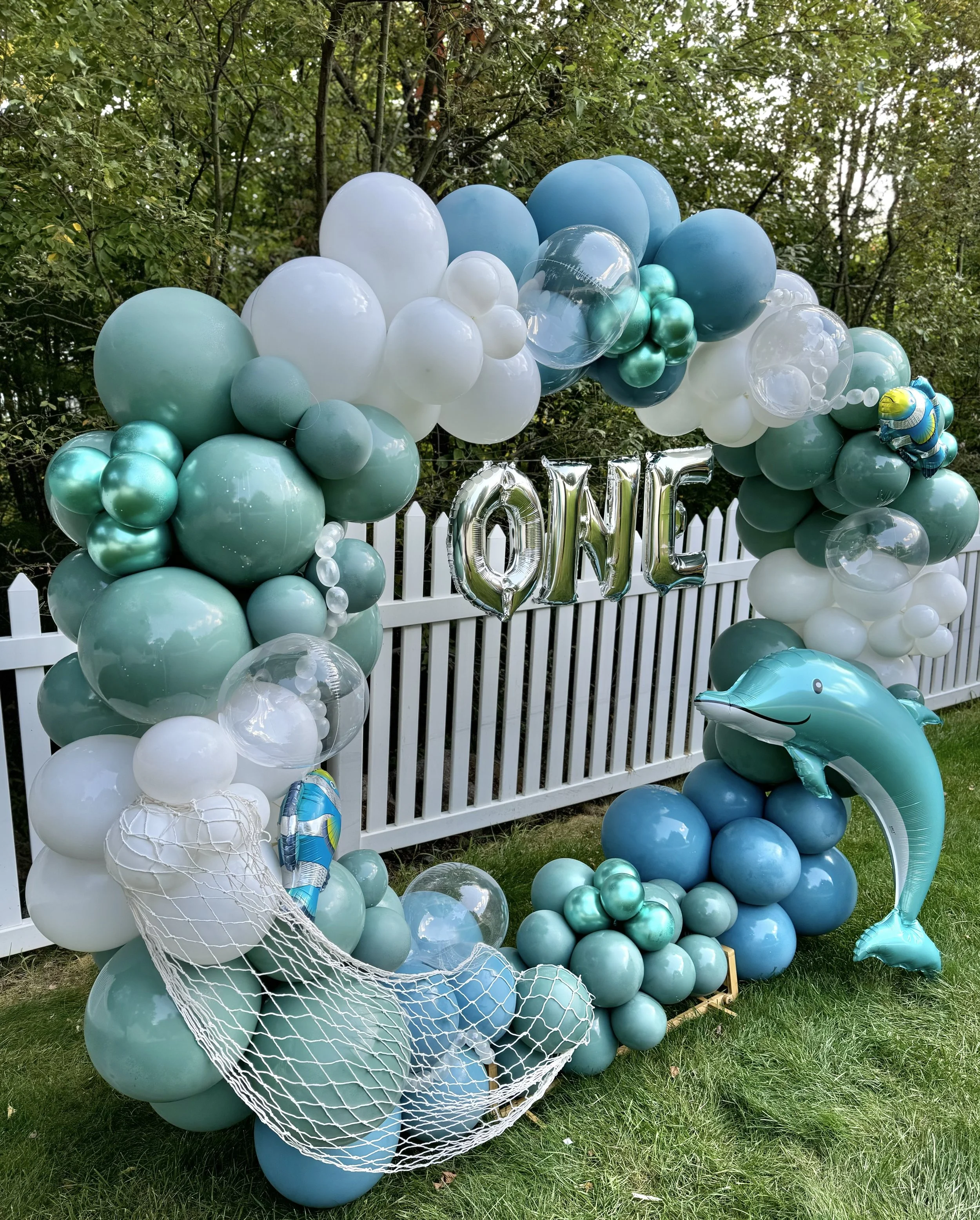 A balloon arrangement for a first birthday celebration featuring a teal, white, and clear color scheme. It includes a balloon arch, a bubble-shaped balloon, a dolphin-shaped balloon, and balloons spelling out 'ONE' in metallic silver letters, set aga