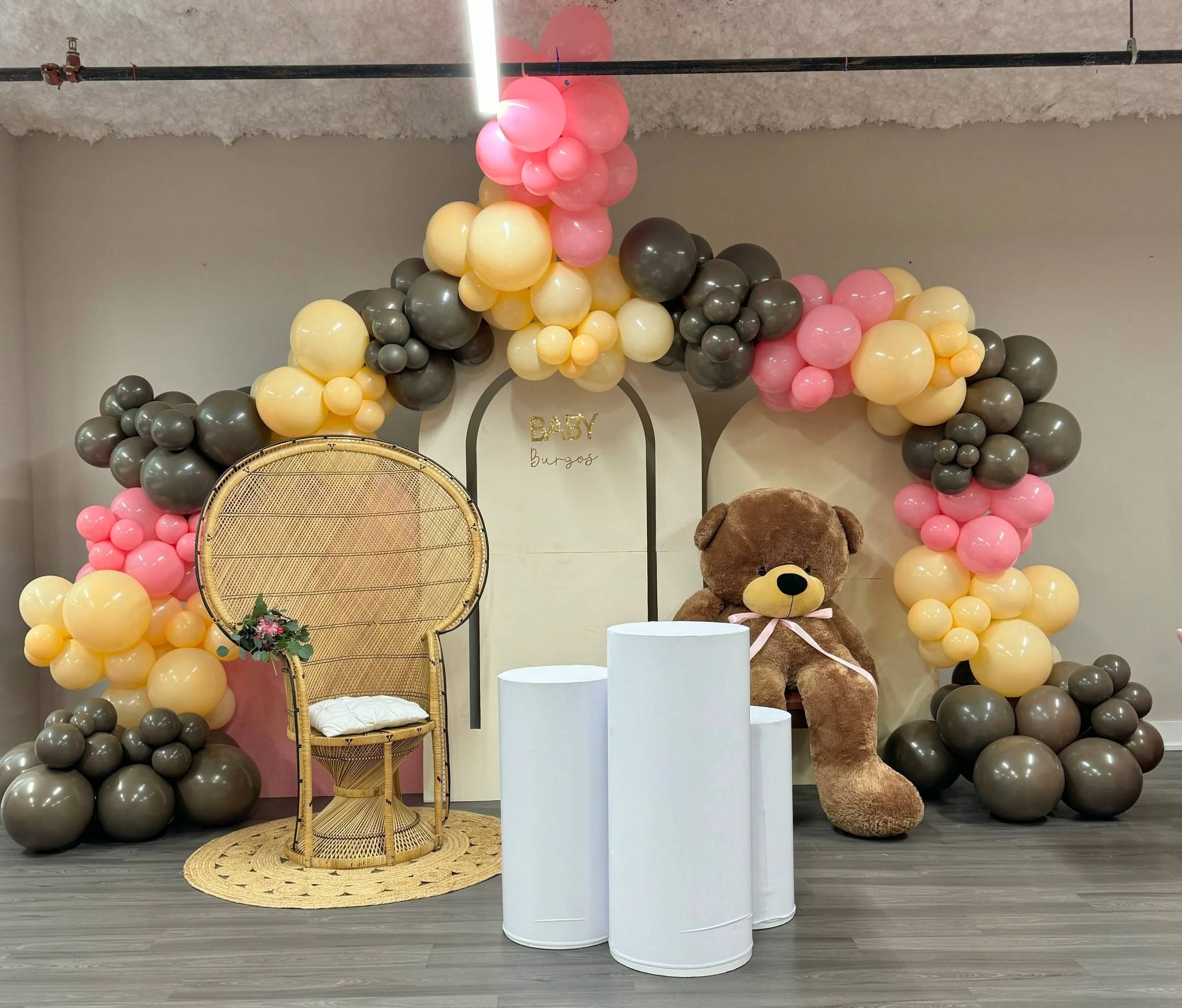 Decorative baby shower backdrop with balloon arch in pink, yellow, and gray, a large teddy bear sitting on a small chair, white cylindrical pedestals, and a woven chair with a floral arrangement.