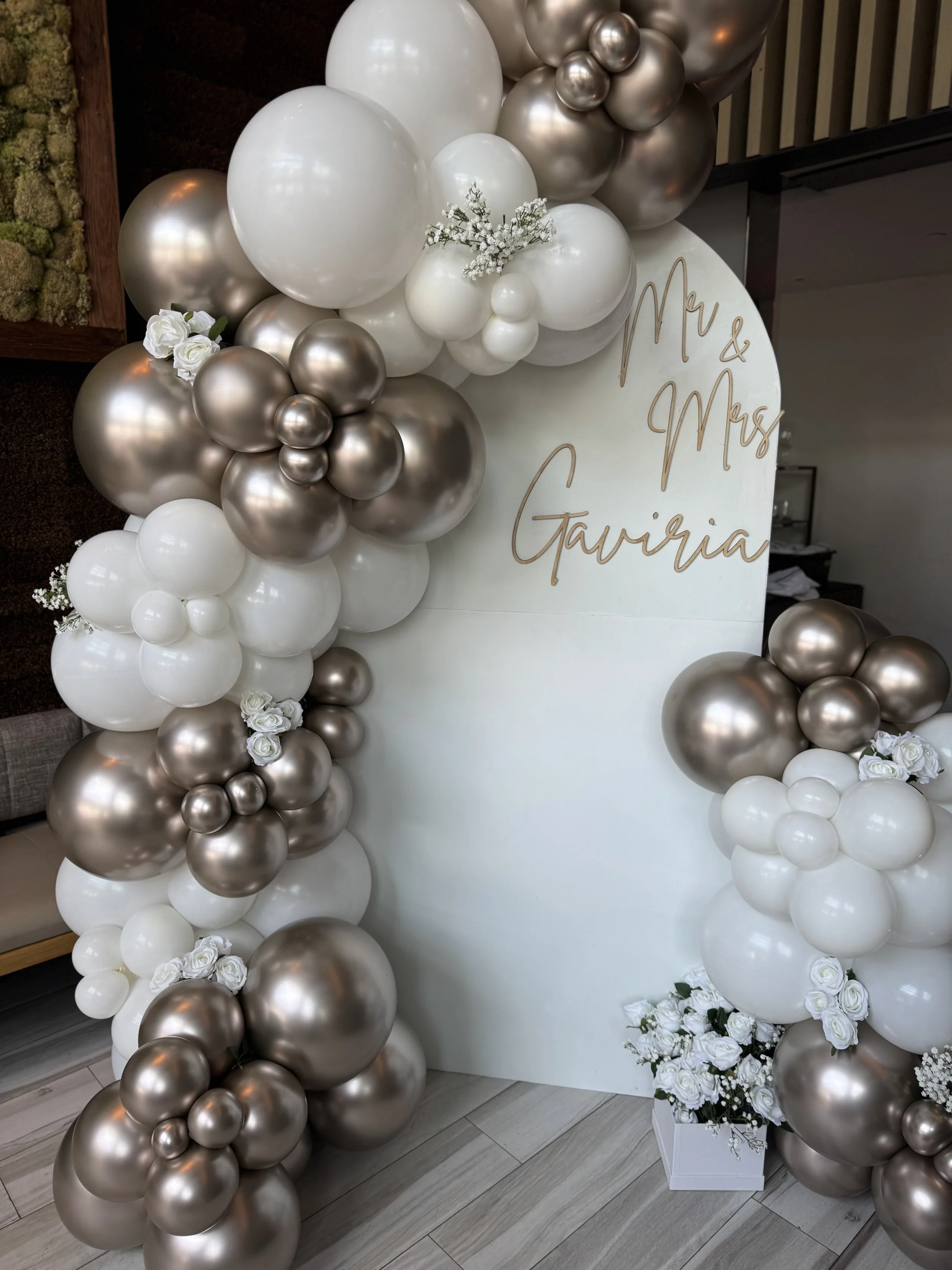 Decorative balloon arch with white, champagne, and metallic balloons, along with white roses and a white backdrop with gold handwritten text reading 'Mr & Mrs Gaviaria'.