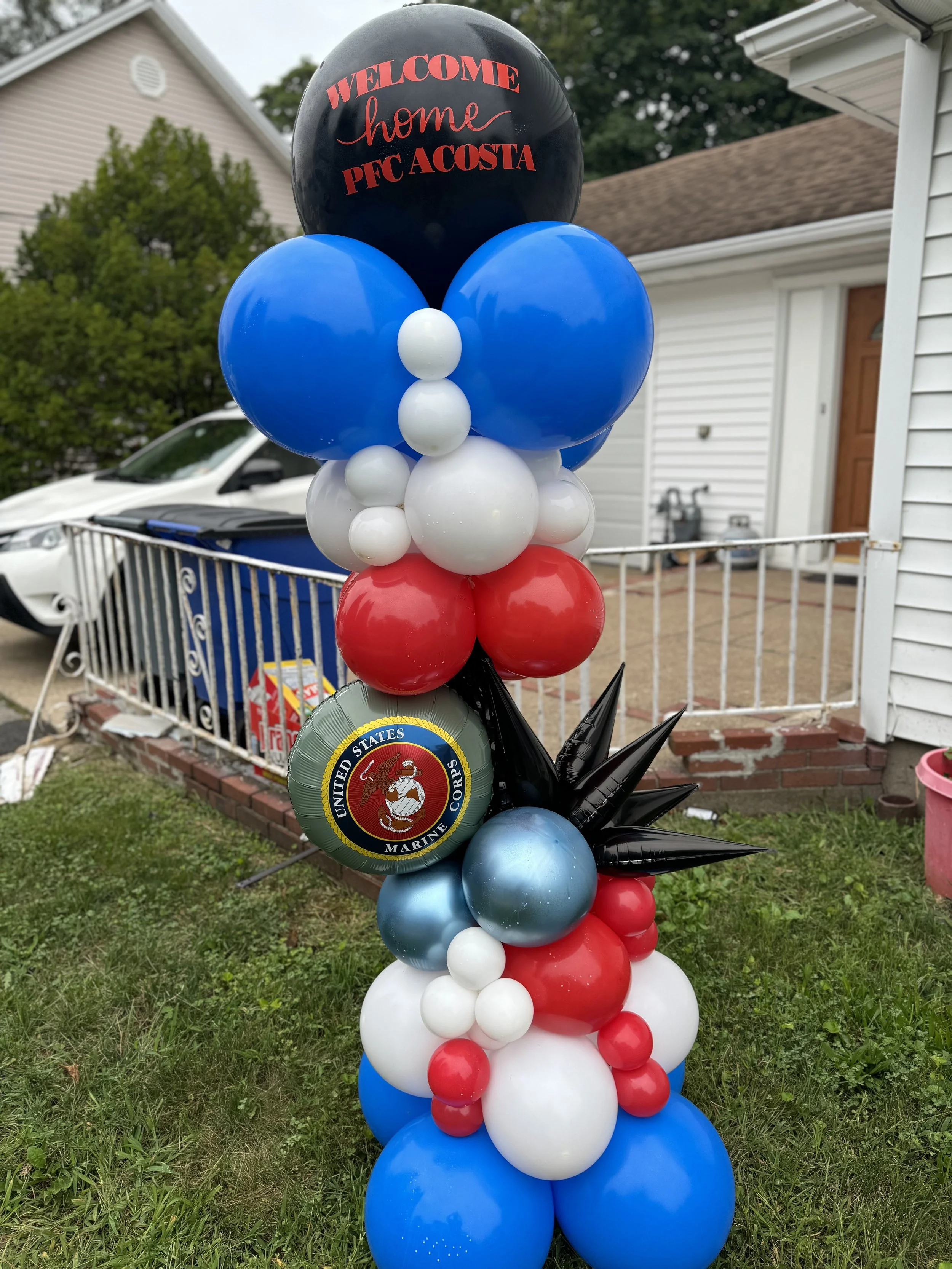 Patriotic balloon arrangement with red, white, blue, and black balloons, including a United States Marine Corps emblem, in a residential yard.