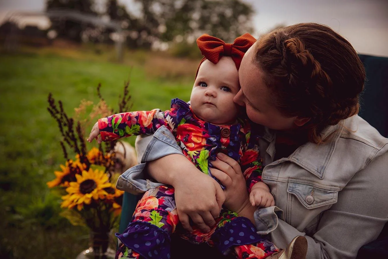 A woman with red hair holding a baby girl outdoors, close to sunset. The baby wears a floral dress with a red bow headband, and there is a bouquet of sunflowers in the background.