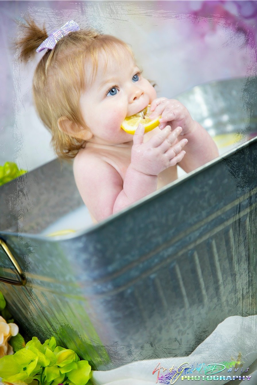 A young girl with red hair and blue eyes is taking a bath, holding a lemon slice and biting into it.