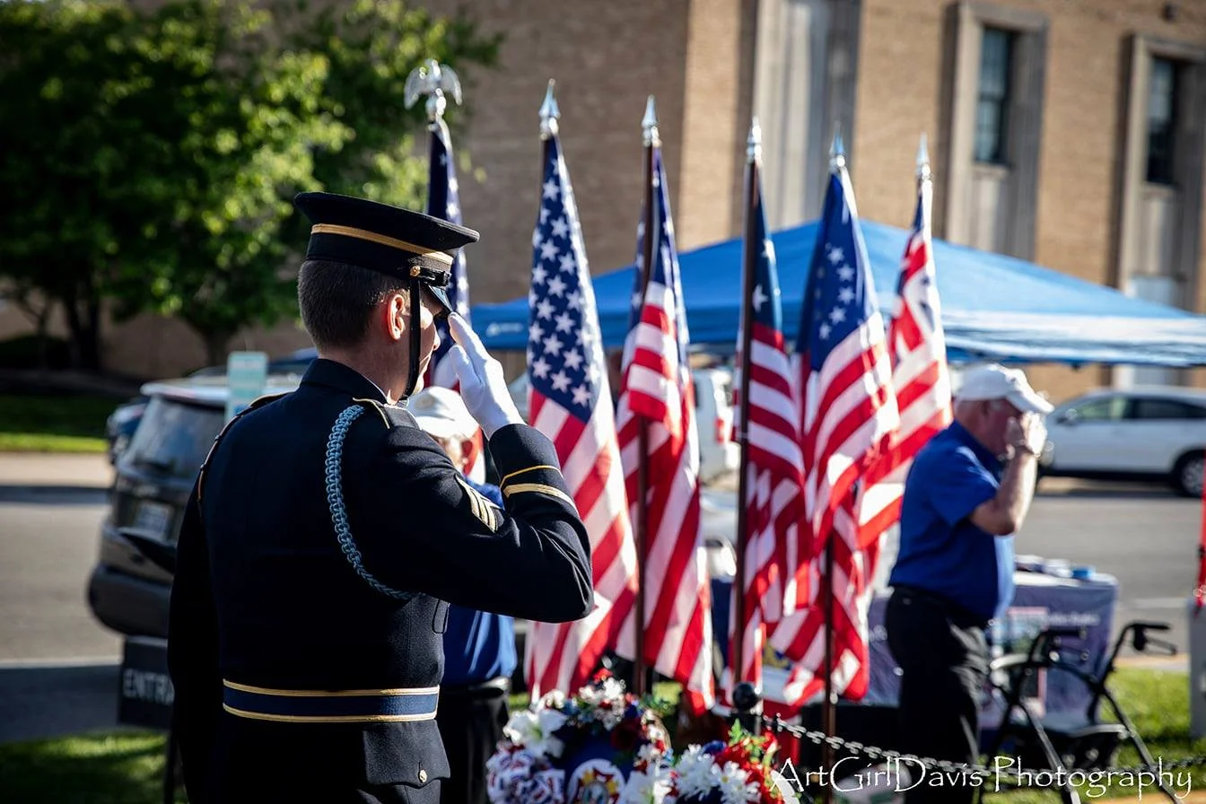A military honor guard salutes at a patriotic memorial event with American flags and floral arrangements.