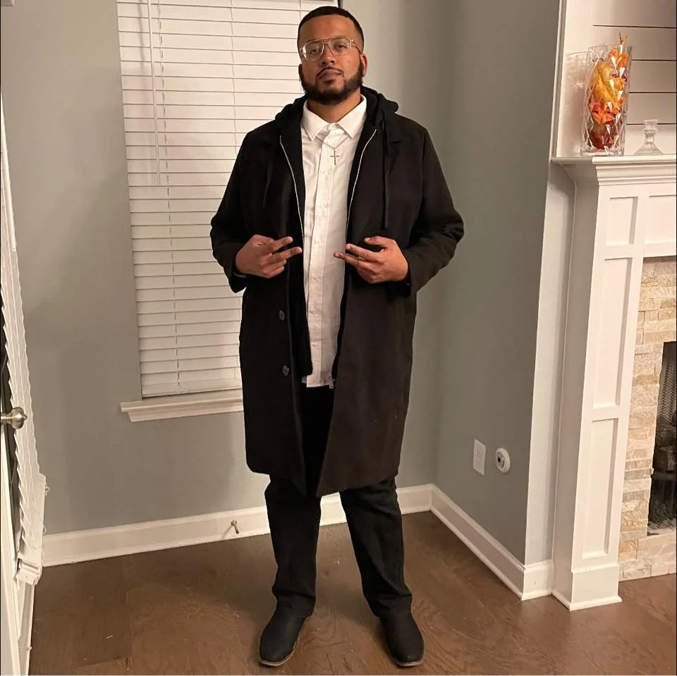 Man wearing black coat and glasses posing indoors next to a fireplace.
