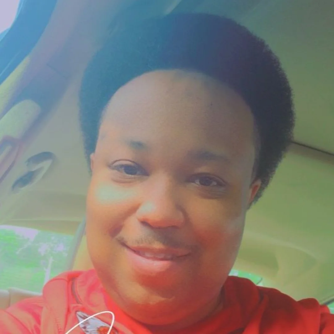 Person smiling inside a car, wearing a red shirt, with short curly hair.