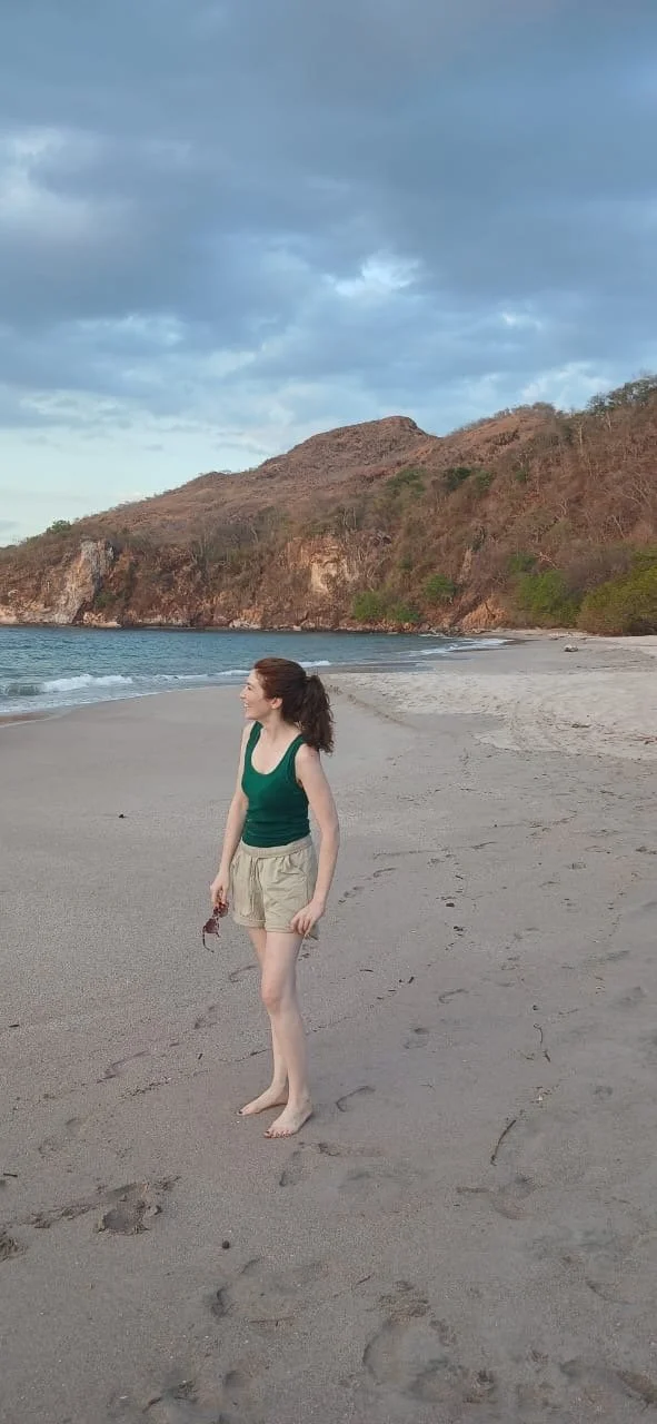 Woman standing barefoot on the sandy beach with hills and cloudy sky in the background.