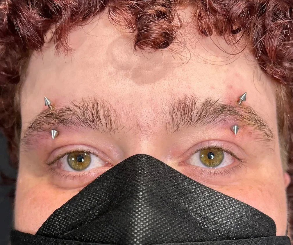 a man with a matching set of eyebrow piercings performed with implant grade titanium curved barbells by a professional body piercer in a professional body piercing studio. 14g curved barbells with spikes in a set of paired eyebrow piercings on a guy.