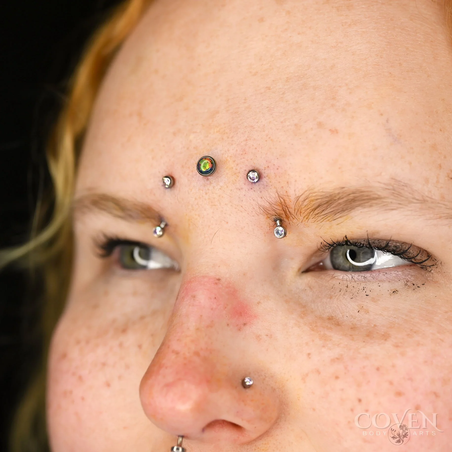 Inner eyebrow piercings on a person who also has a third eye dermal and a nostril piercing. Eyebrow piercings performed with implant grade titanium curved barbells from NeoMetal Body Jewelry and pierced by Dee at Coven Body Arts, a professional body 