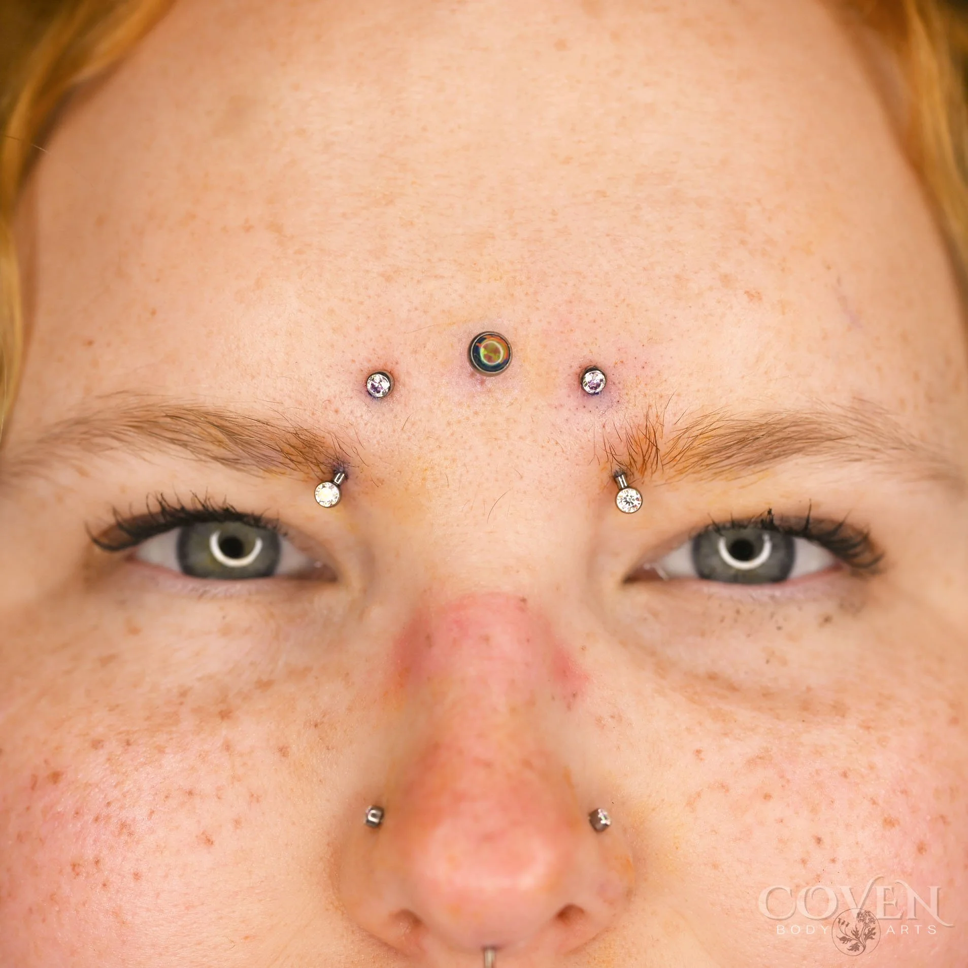 Inner eyebrow piercings on a person who also has a third eye dermal and paired nostril piercing. Eyebrow piercings performed with implant grade titanium curved barbells from NeoMetal Body Jewelry and pierced by Dee at Coven Body Arts, a professional 
