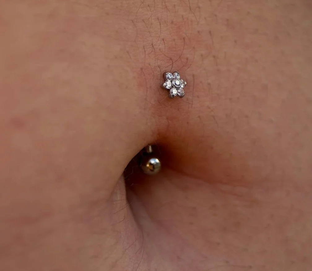 closeup of a fresh navel piercing performed with an implant grade titanium 14g curved barbell with a gemmed flower on the top. side view of a navel piercing on a women with a flower barbell.  a navel piercing featuring jewelry with a sparkly flower t