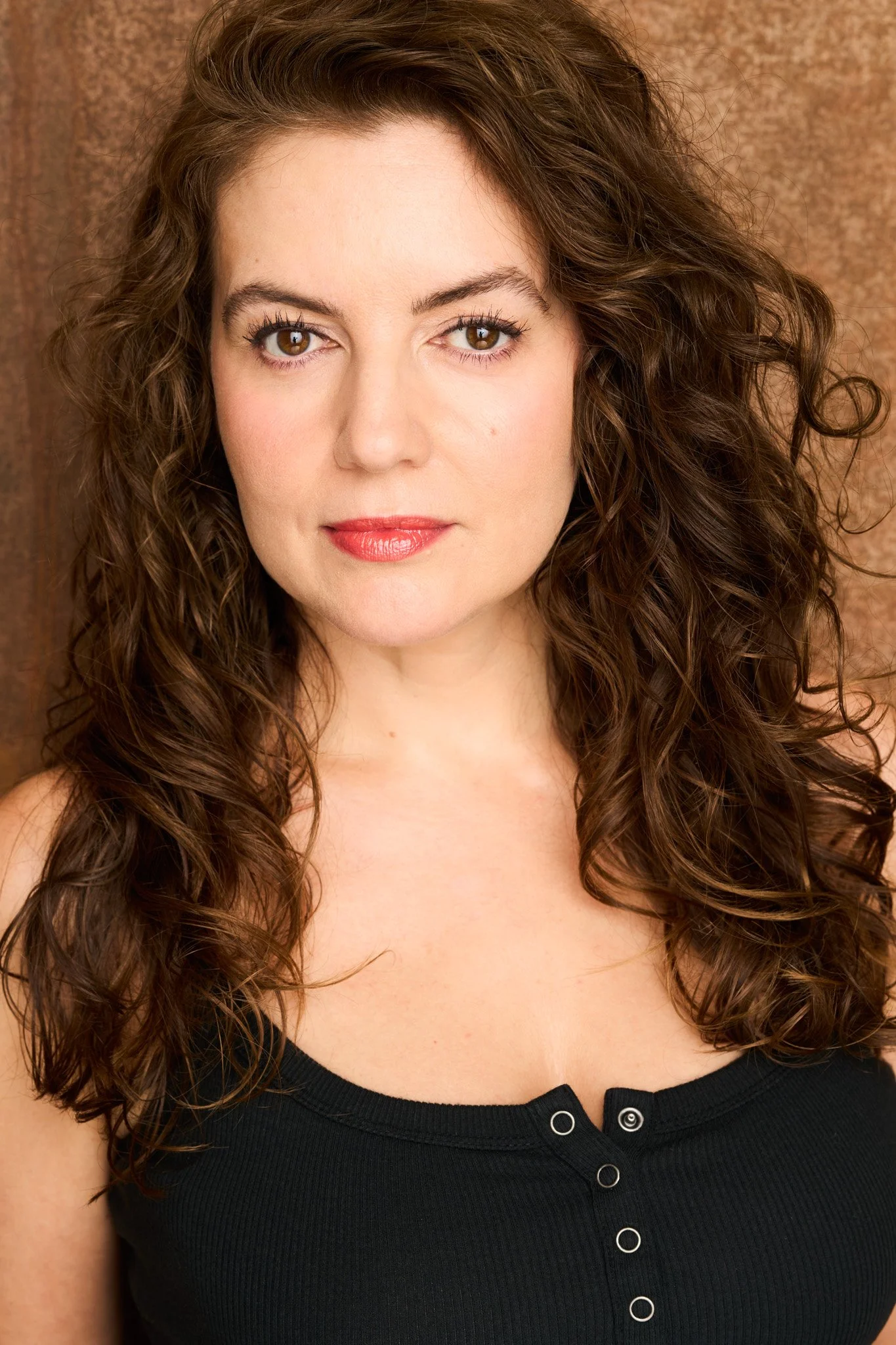 Dramatic headshot of actress Lorena Fernández with long curly hair and a black top against a textured brown background.