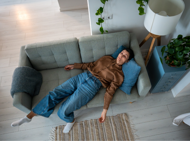A woman lying on a gray sofa with her eyes closed, wearing a brown silk blouse and blue jeans, in a modern living room with light wooden floors and green plants.