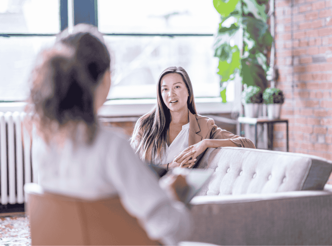 Two women having a conversation in a bright, modern office or lounge with large windows, a brick wall, and plants, one woman is speaking while the other listens.