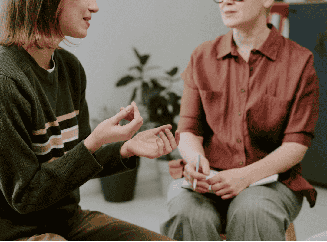 Two women engaging in conversation, one explaining with hand gestures and the other taking notes with a pen, in a therapy or counseling session setting.