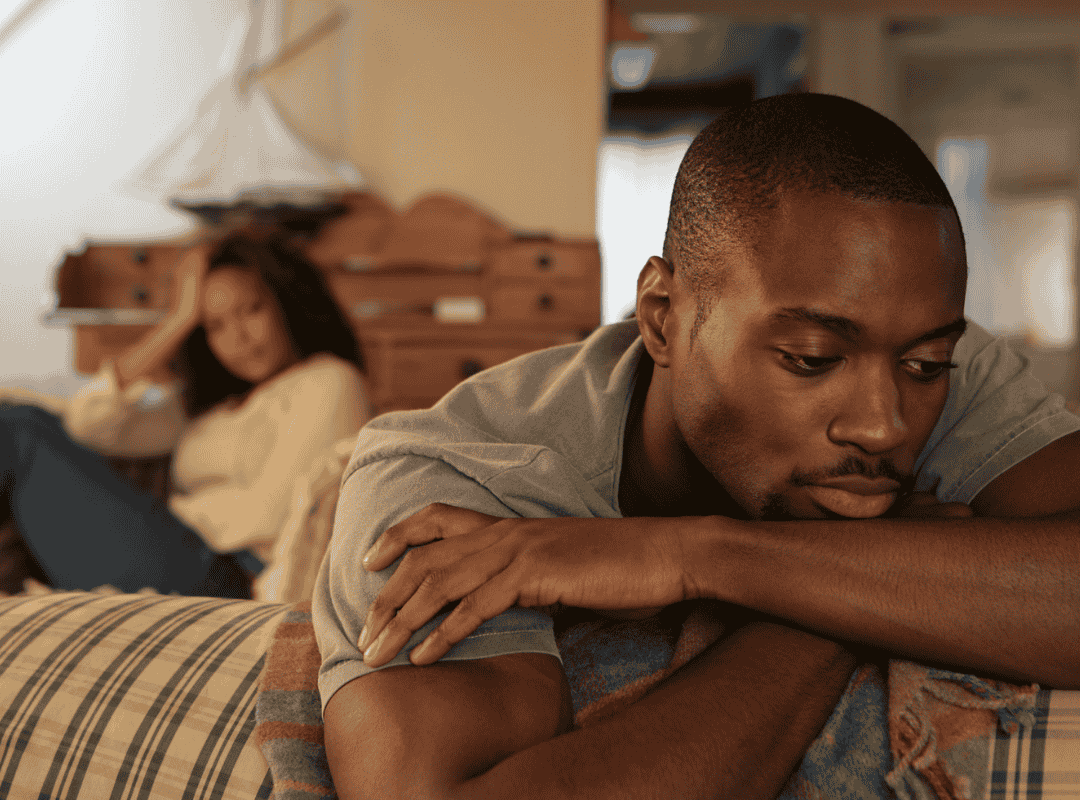 A man lying on his stomach on a bed, looking sad or contemplative, with a woman in the background sitting on the bed.