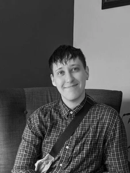 A person with short dark hair and a slight smile, sitting on a cushioned chair indoors, wearing a plaid shirt and carrying a shoulder bag.