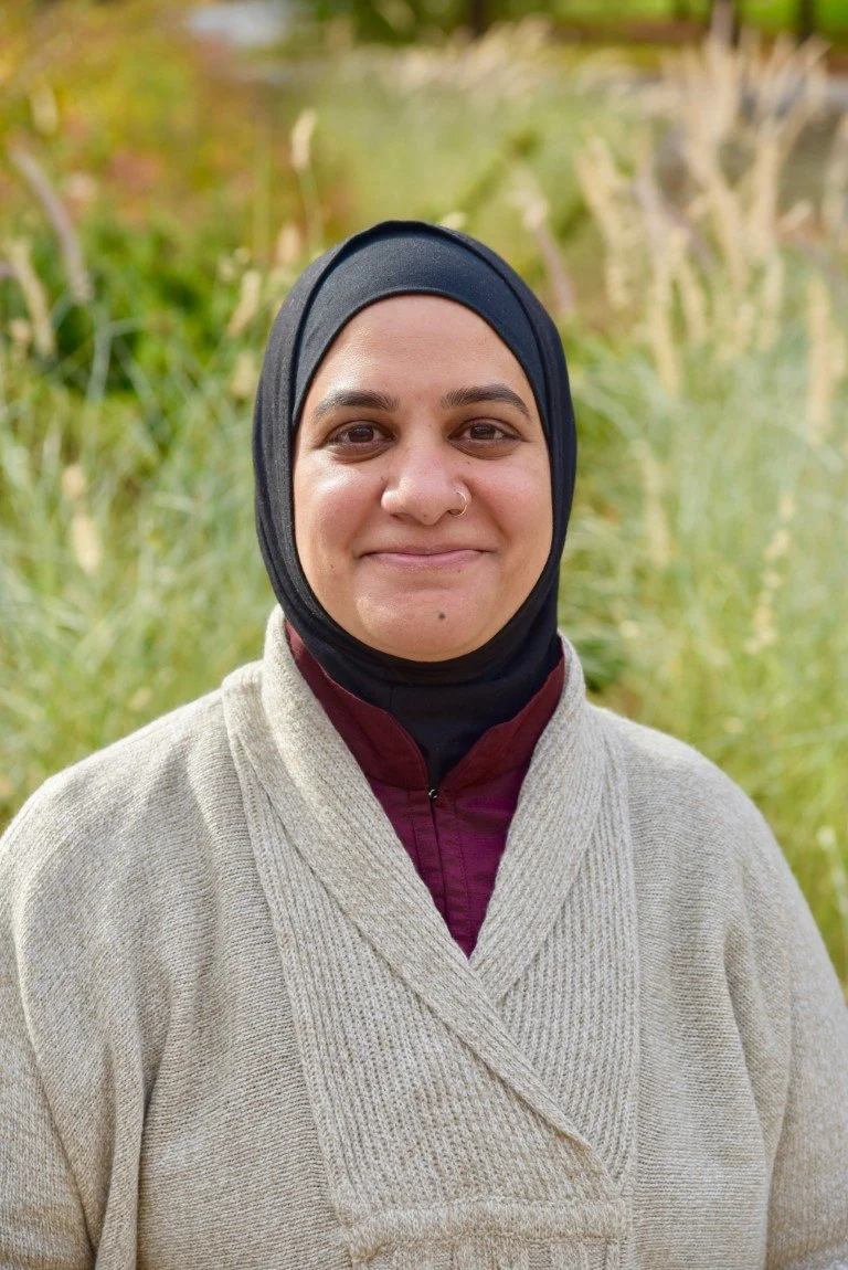female Muslim therapist with a black headscarf and beige cardigan standing in a field smiling at the camera