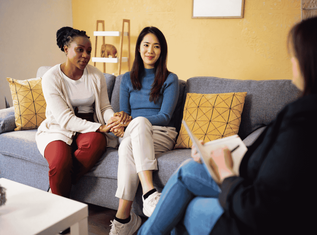 Two women sitting on a gray couch holding hands and talking, while a therapist or counselor taking notes listens during a therapy session in a cozy room with yellow walls and geometric-patterned pillows.