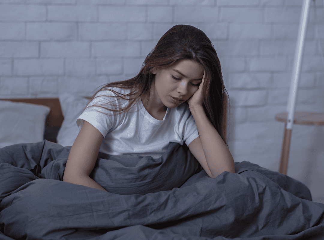A woman sitting on a bed with a gray blanket, looking distressed and holding her head.