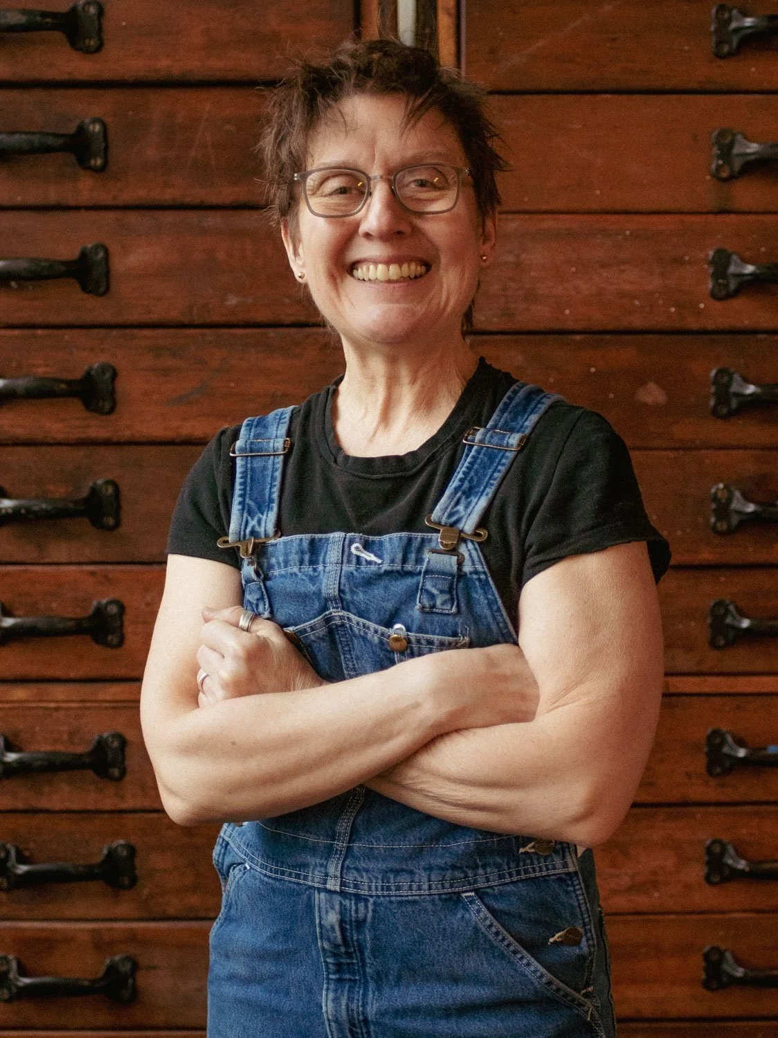 Portrait of smiling Karen Beckwith, Tamarind certified Master Printmaker and professional Fine Art Printmaker, wearing blue overalls and standing in front of wooden print drawers.