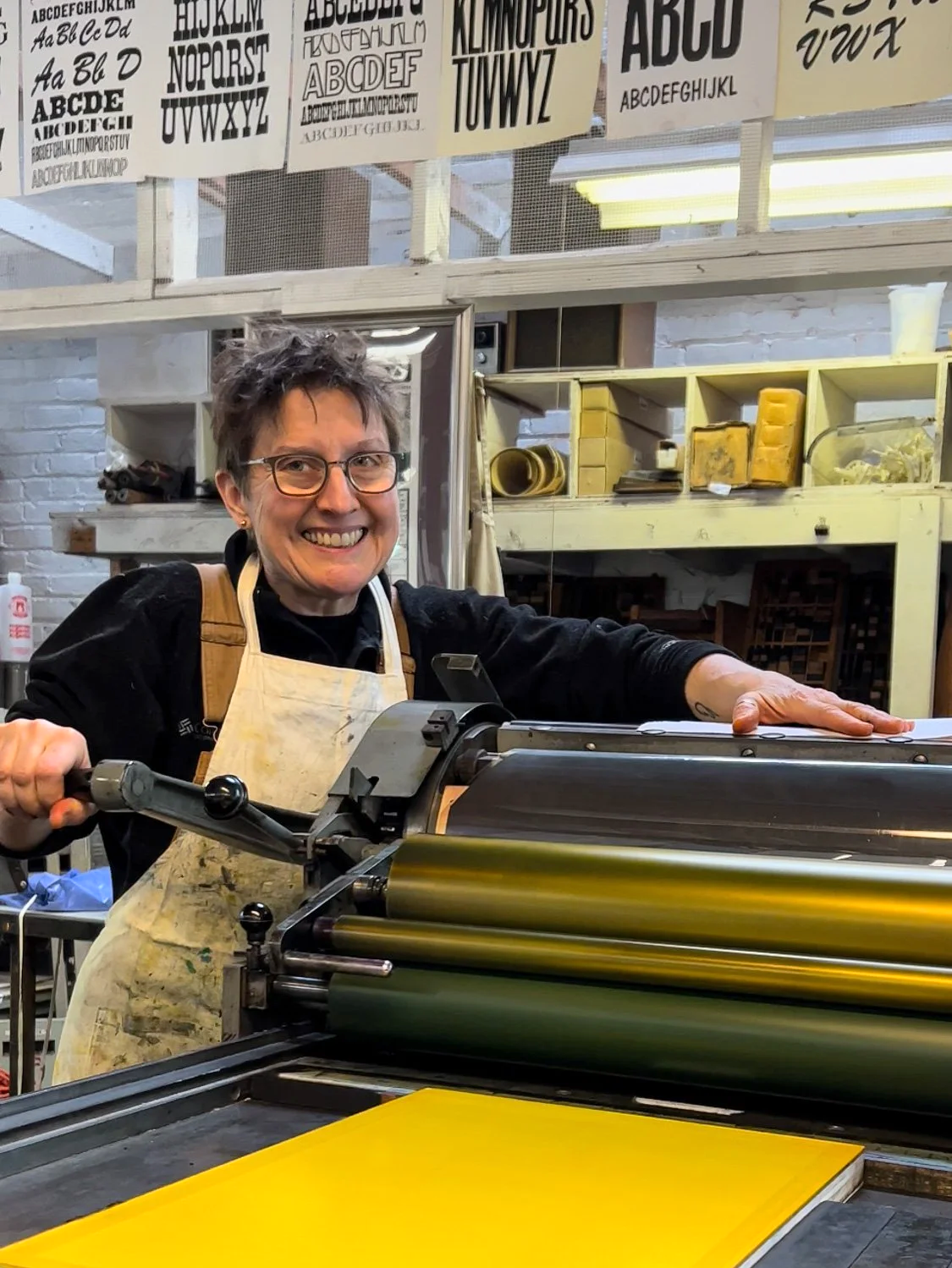 Karen Beckwith, Cleveland printmaker, using a printing press in a workshop. She wears glasses and an apron, with printmaking supplies and equipment in the background.