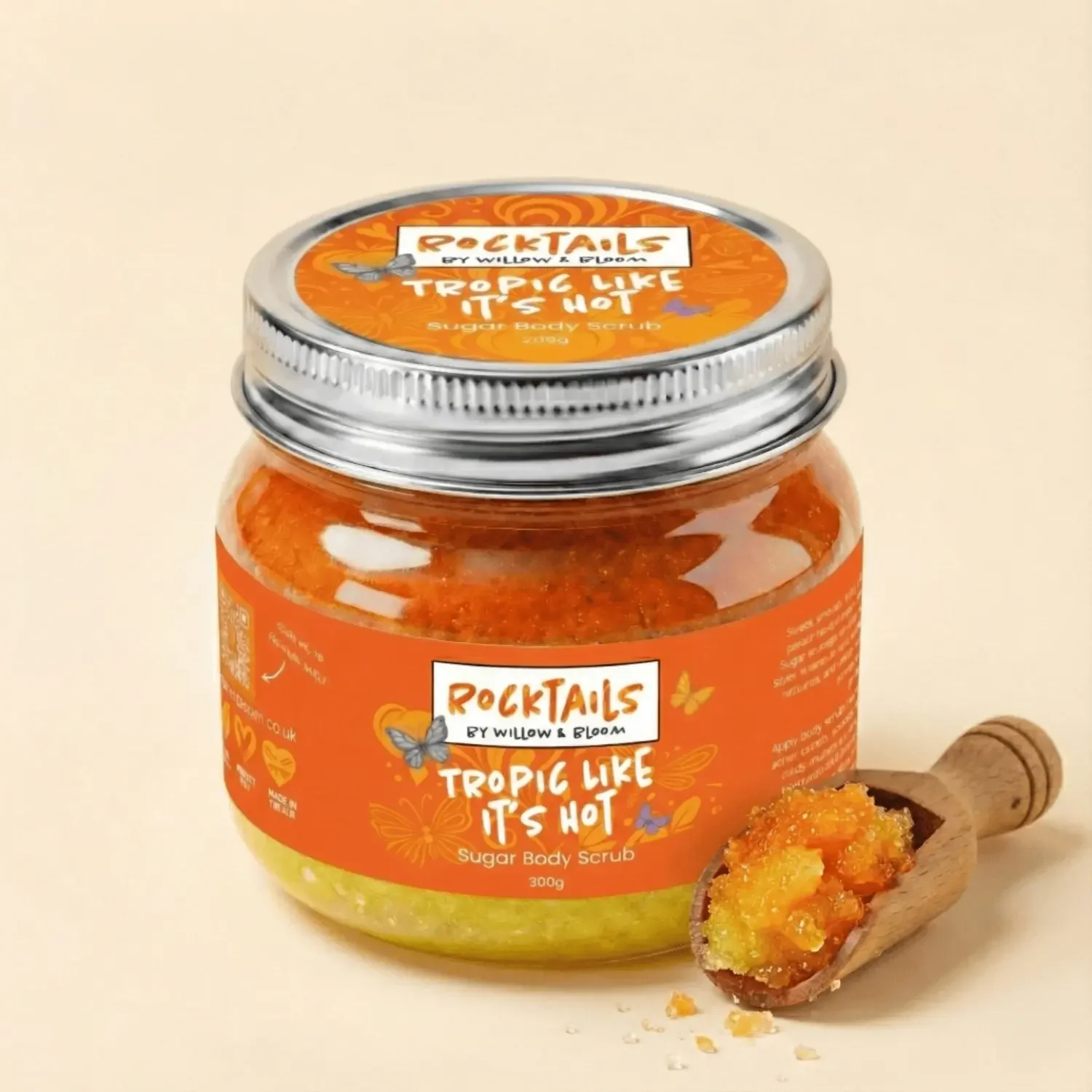 A jar of orange sugar body scrub labeled 'Rocktails by Willow & Bloom, Tropical Like It's Hot,' with a wooden scoop beside it filled with the scrub.