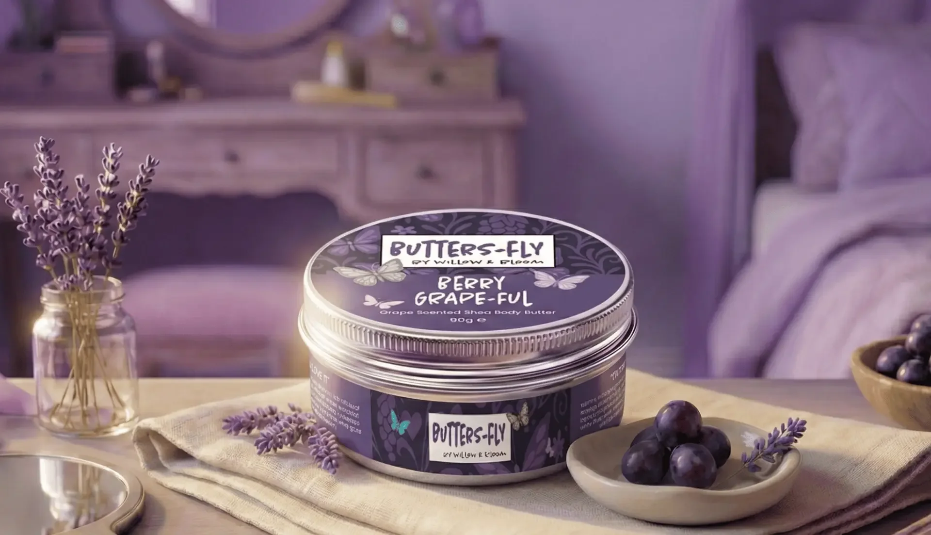 A jar of butters-fly body butter with a purple lavender theme, placed on a beige cloth next to a small dish of grapes and sprigs of lavender, in a softly lit bedroom setting.
