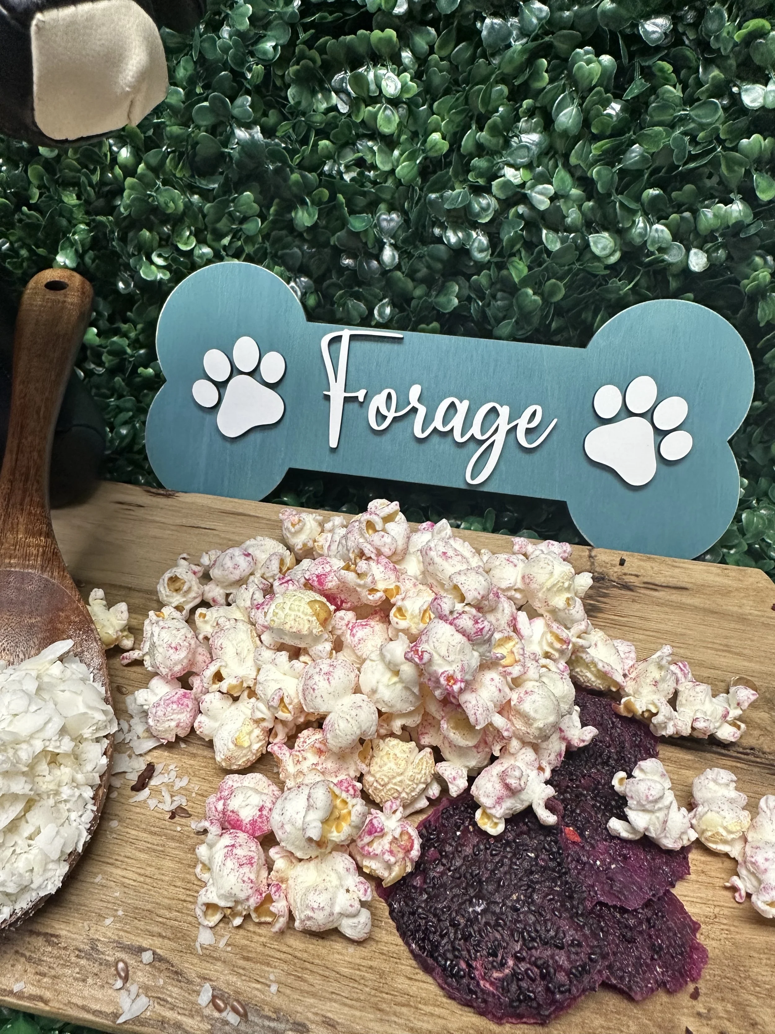 Popcorn with pink seasoning and purple chips on a wooden surface, surrounded by greenery background and a dog bone-shaped sign with paw prints.