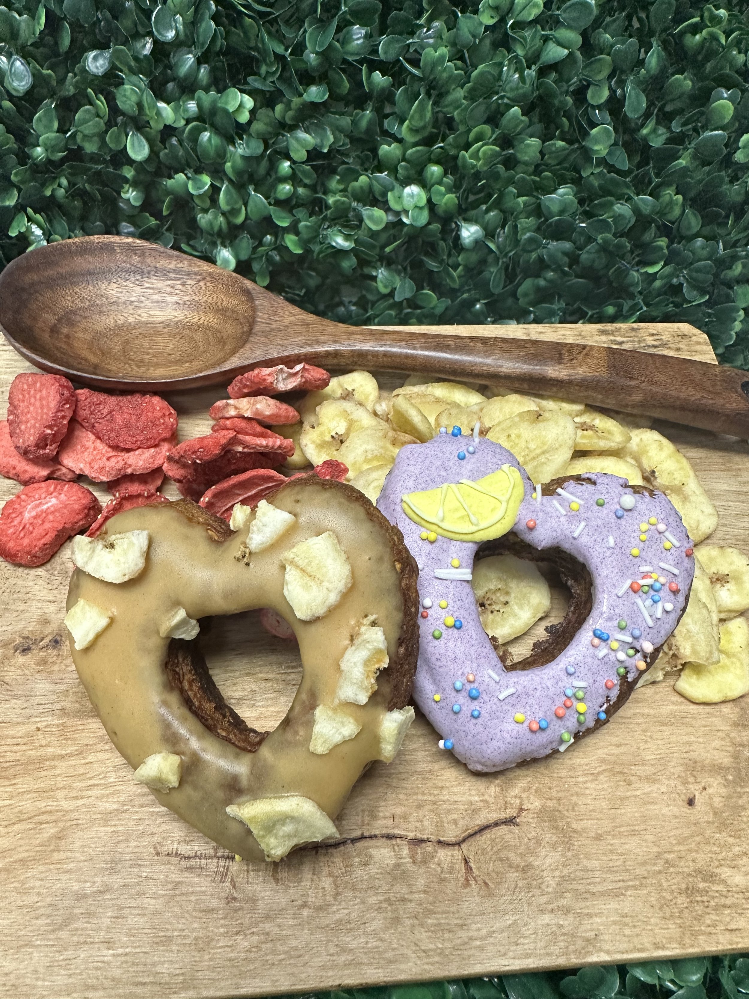 Two heart-shaped donuts on a wooden board, one with caramel glaze and potato chip pieces, the other with purple icing and colorful sprinkles, surrounded by dried fruit slices and a wooden spoon, with a leafy background.