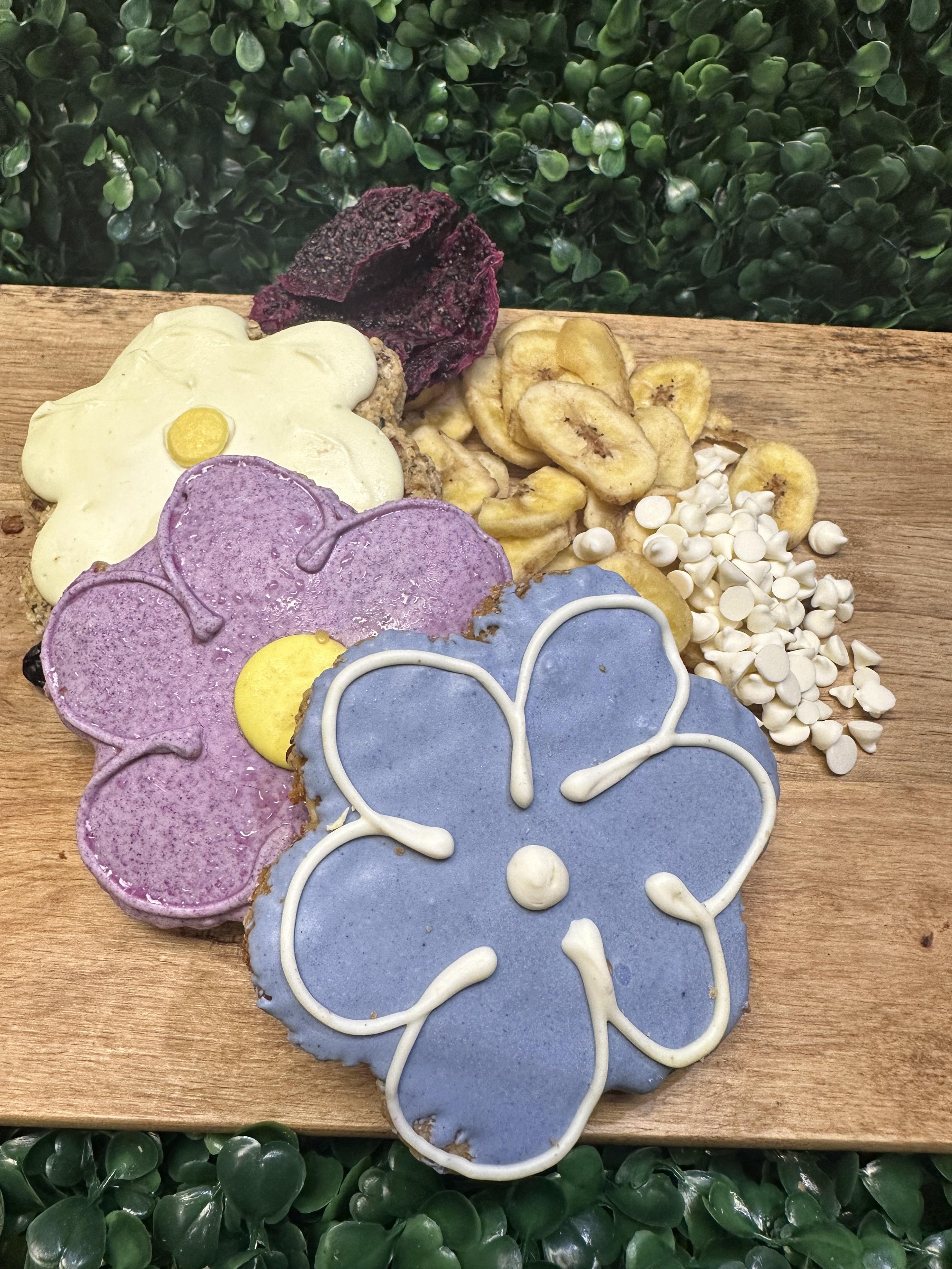 Flower-shaped cookies with colorful icing on a wooden board, surrounded by banana chips, white chocolate chips, and dried fruit slices.