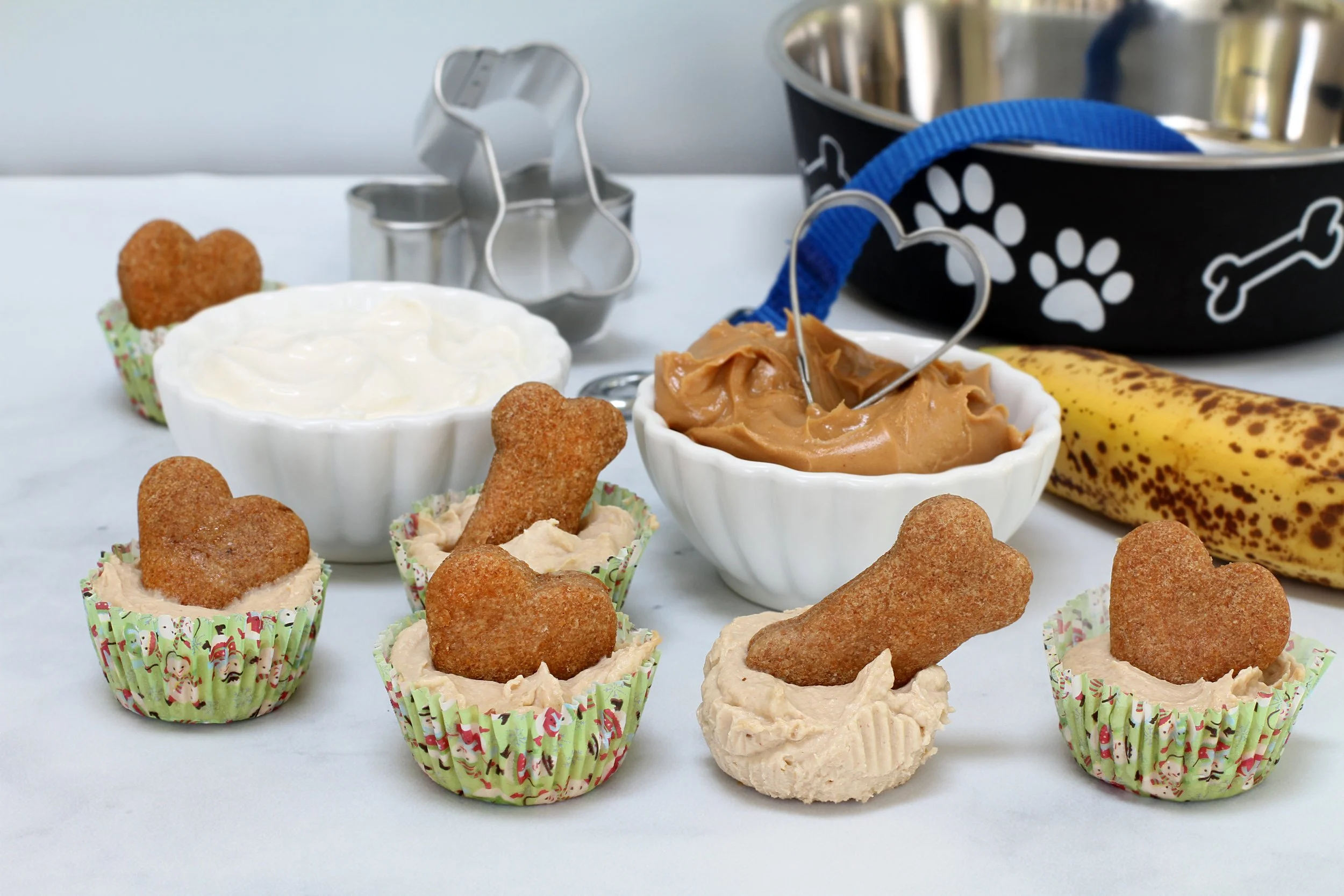 Dog cupcakes with bone-shaped treats, yogurt, peanut butter, banana, and dog-themed accessories on white background.
