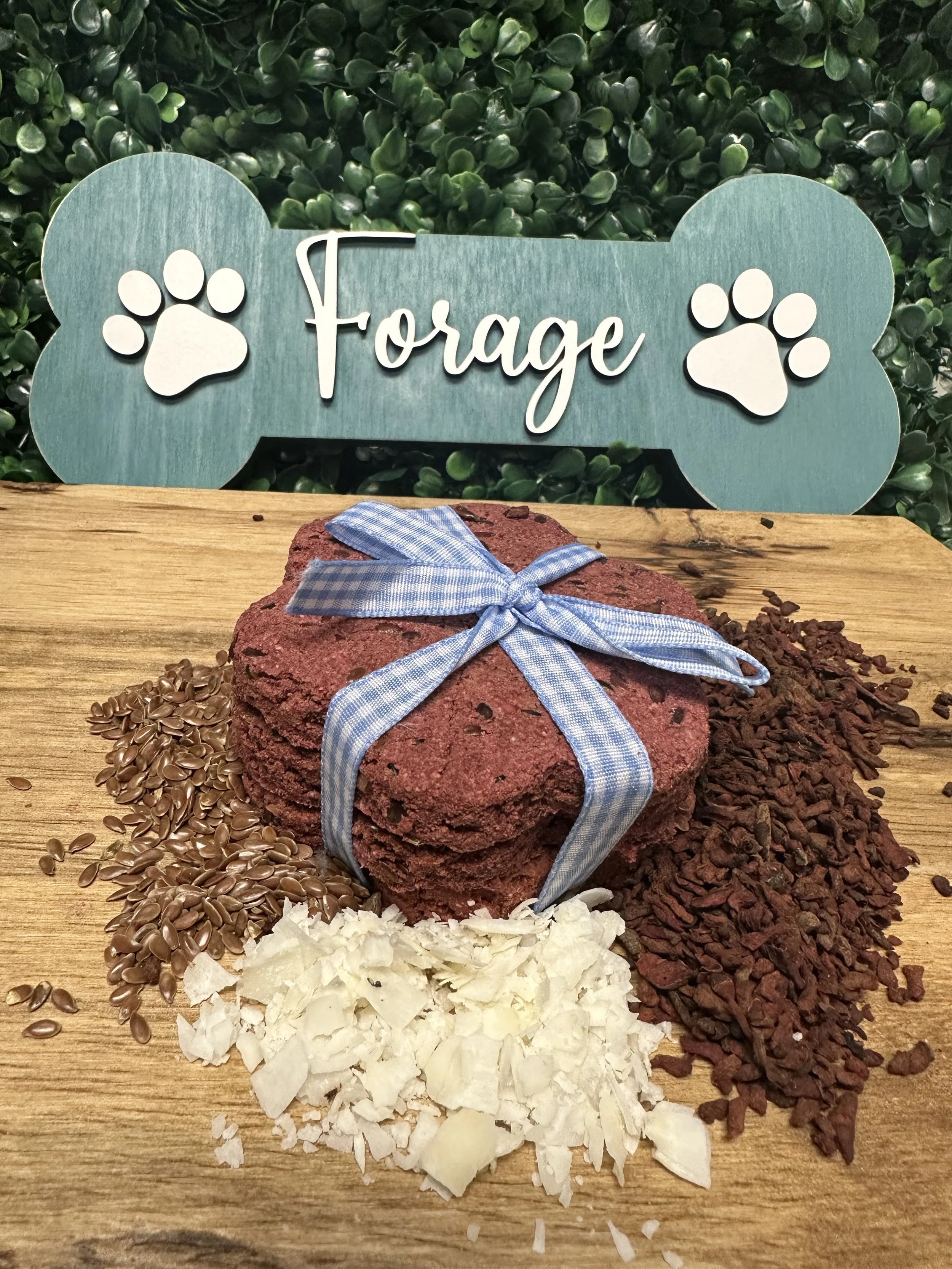 Dog treats tied with a ribbon, surrounded by flaxseeds, coconut flakes, and small dried ingredients on a wooden surface with a green background and bone-shaped sign that reads 'Forage' with paw prints.