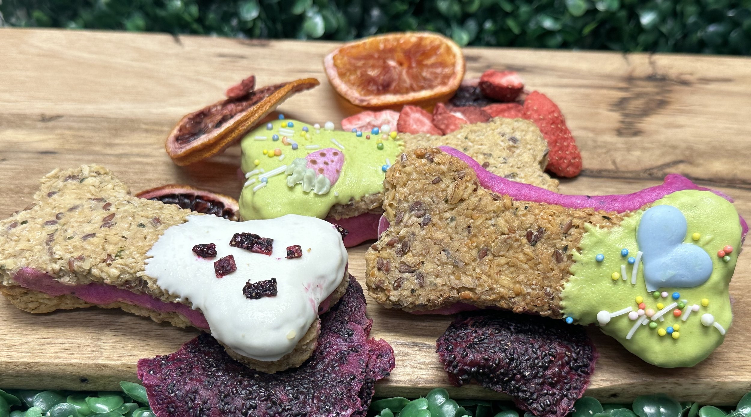 Decorative dog biscuits shaped like bones with various colorful icing and sprinkles, accompanied by dried fruit slices, arranged on a wooden board.