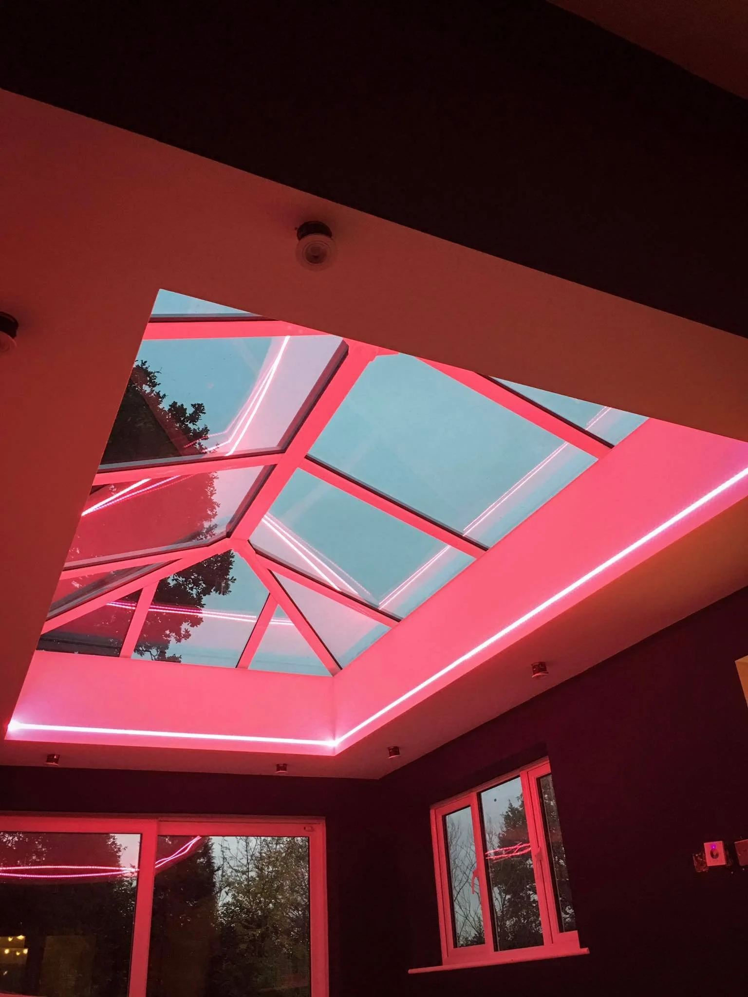 Indoor room with a glass ceiling illuminated with pink neon lights, and large windows showing trees outside.