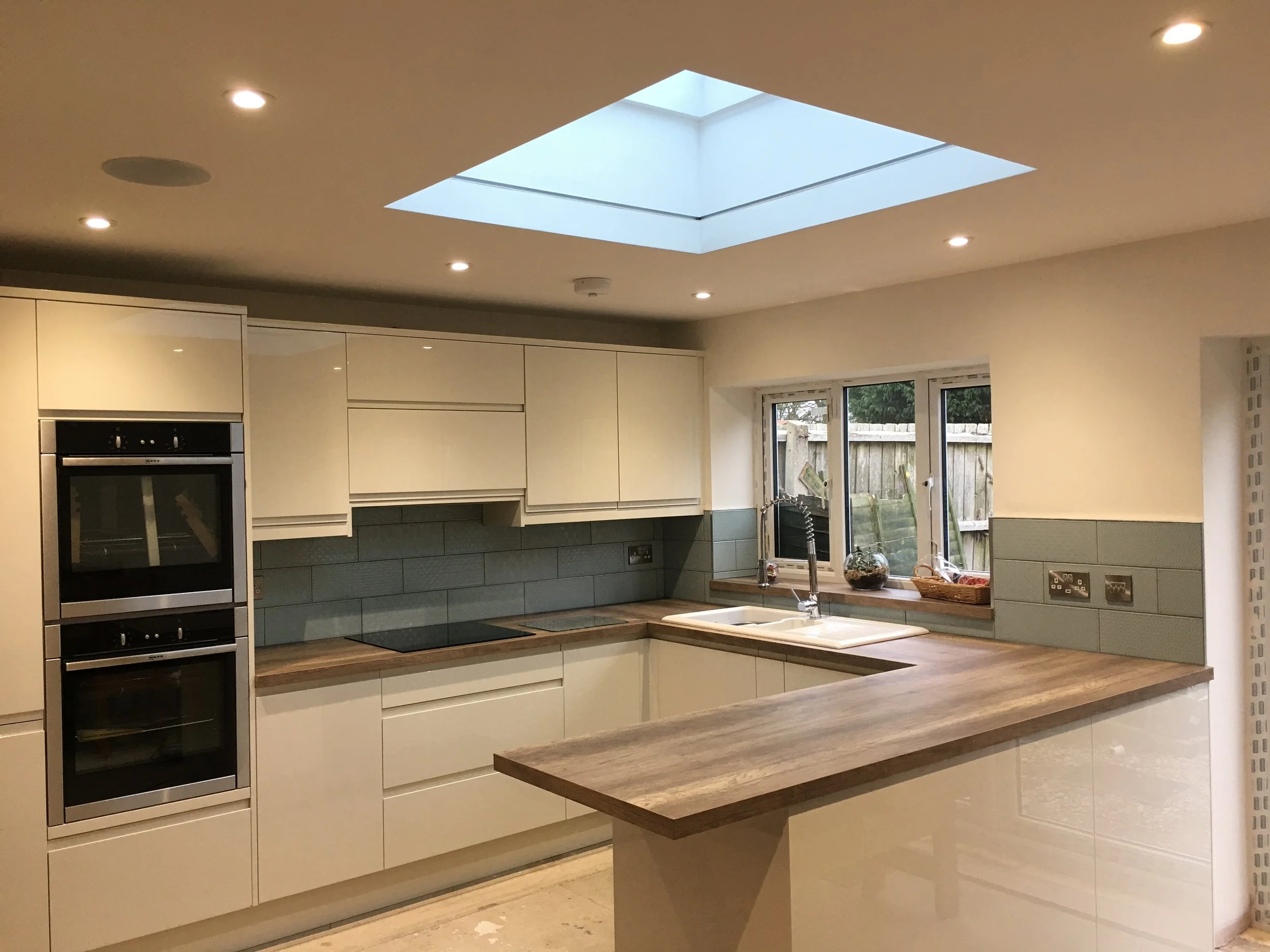Modern kitchen with white cabinetry, wood countertop breakfast bar, double oven, gray tile backsplash, and a window above the sink, illuminated by ceiling lights and skylight.