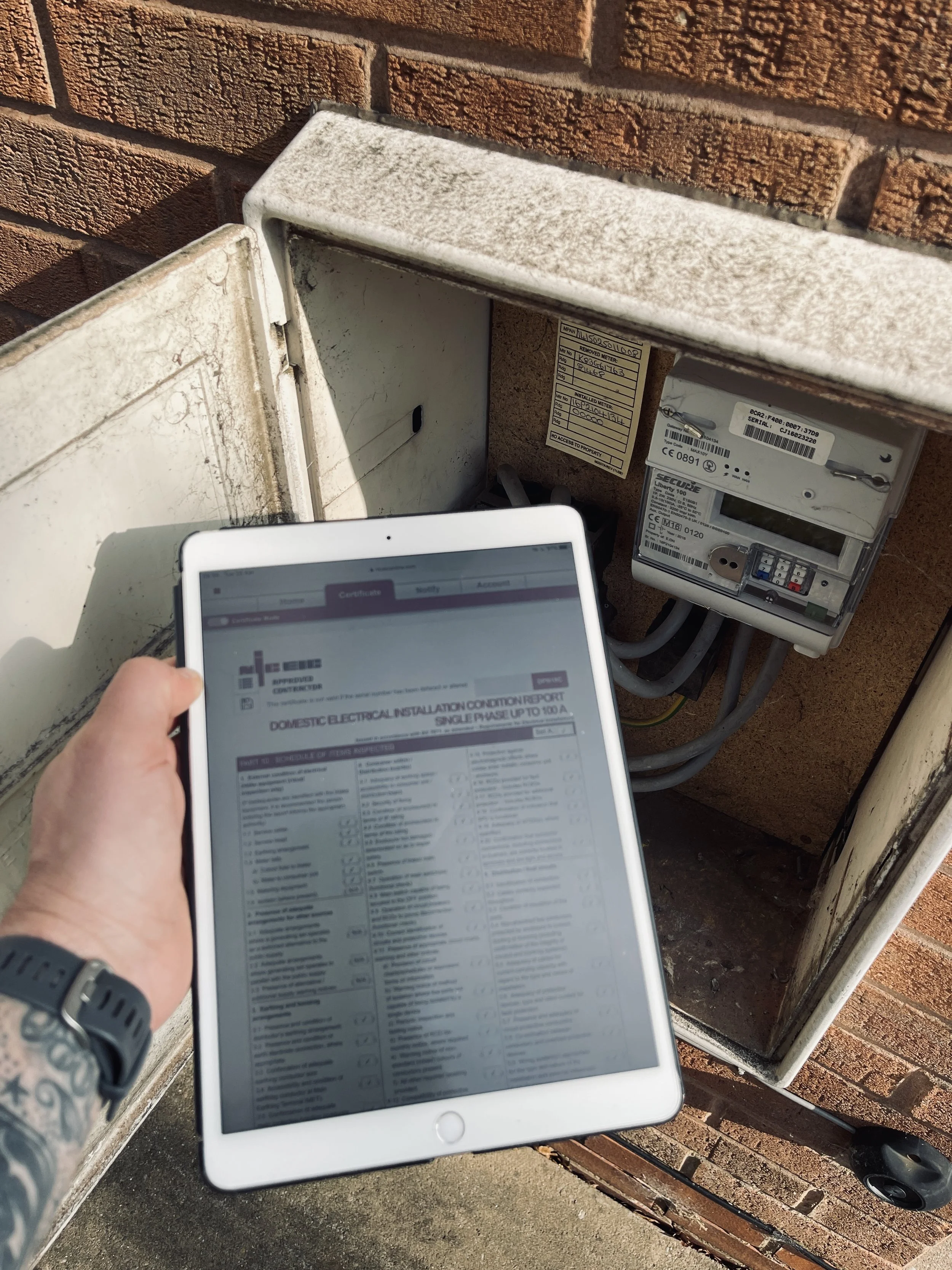 Person holding a tablet in front of an outdoor electrical meter with a brick wall background.