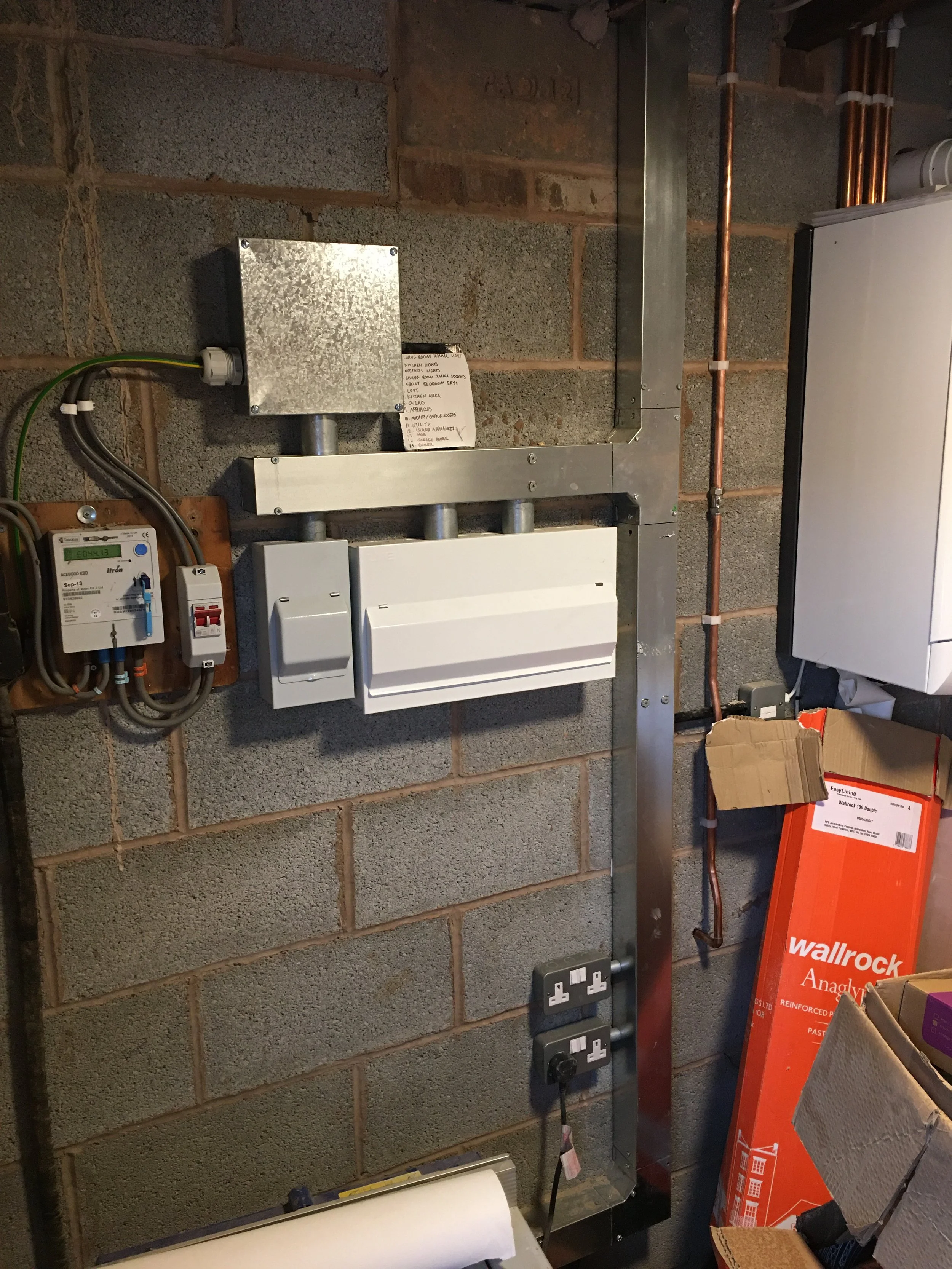 Electrical wiring and conduit on a cinder block wall, including an electrical box, a meter, and a white fuse box with cables and plugs, near boxes and boxes of insulation.